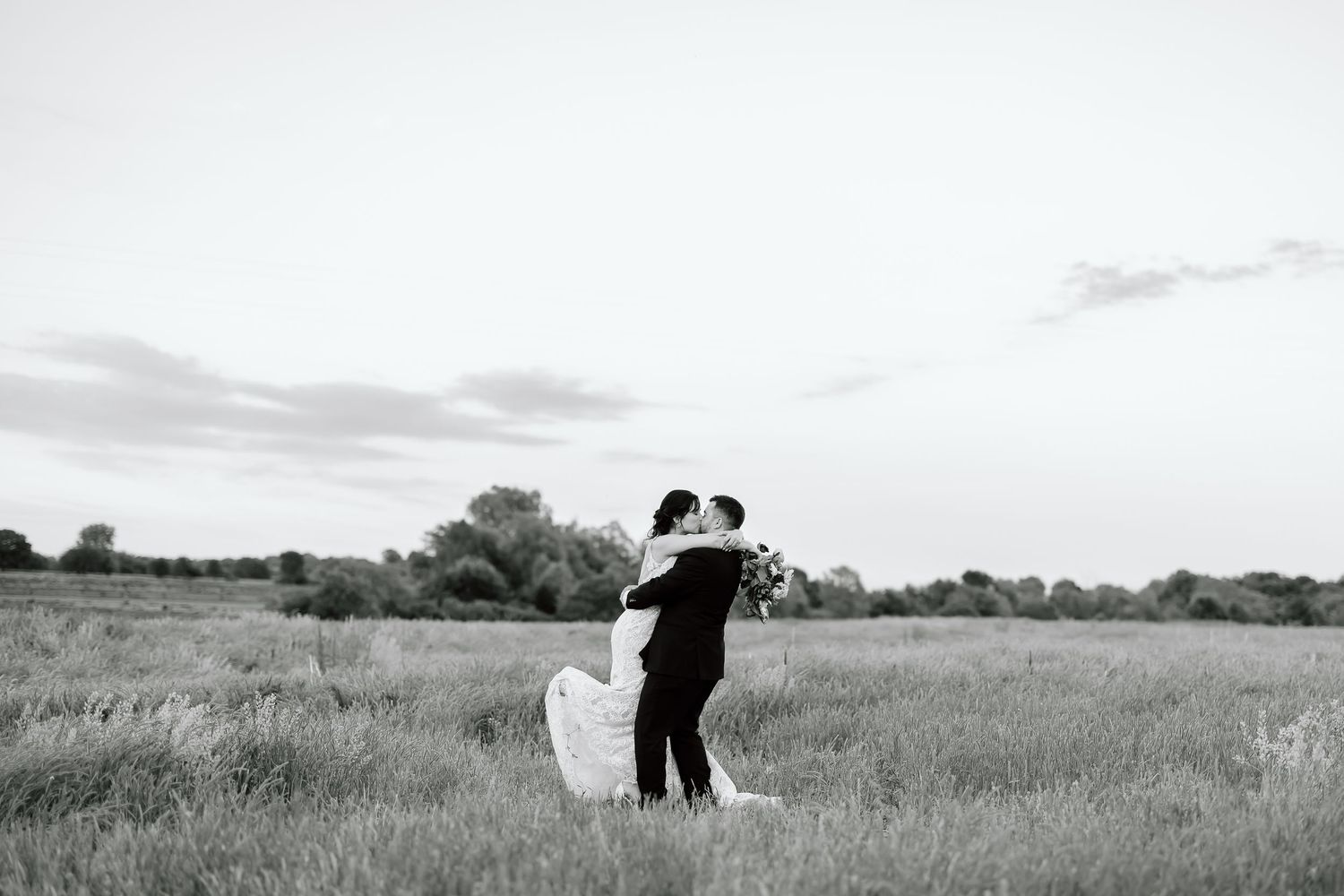A couple shares a loving kiss in a picturesque countryside field during sunset, creating a romantic scene.