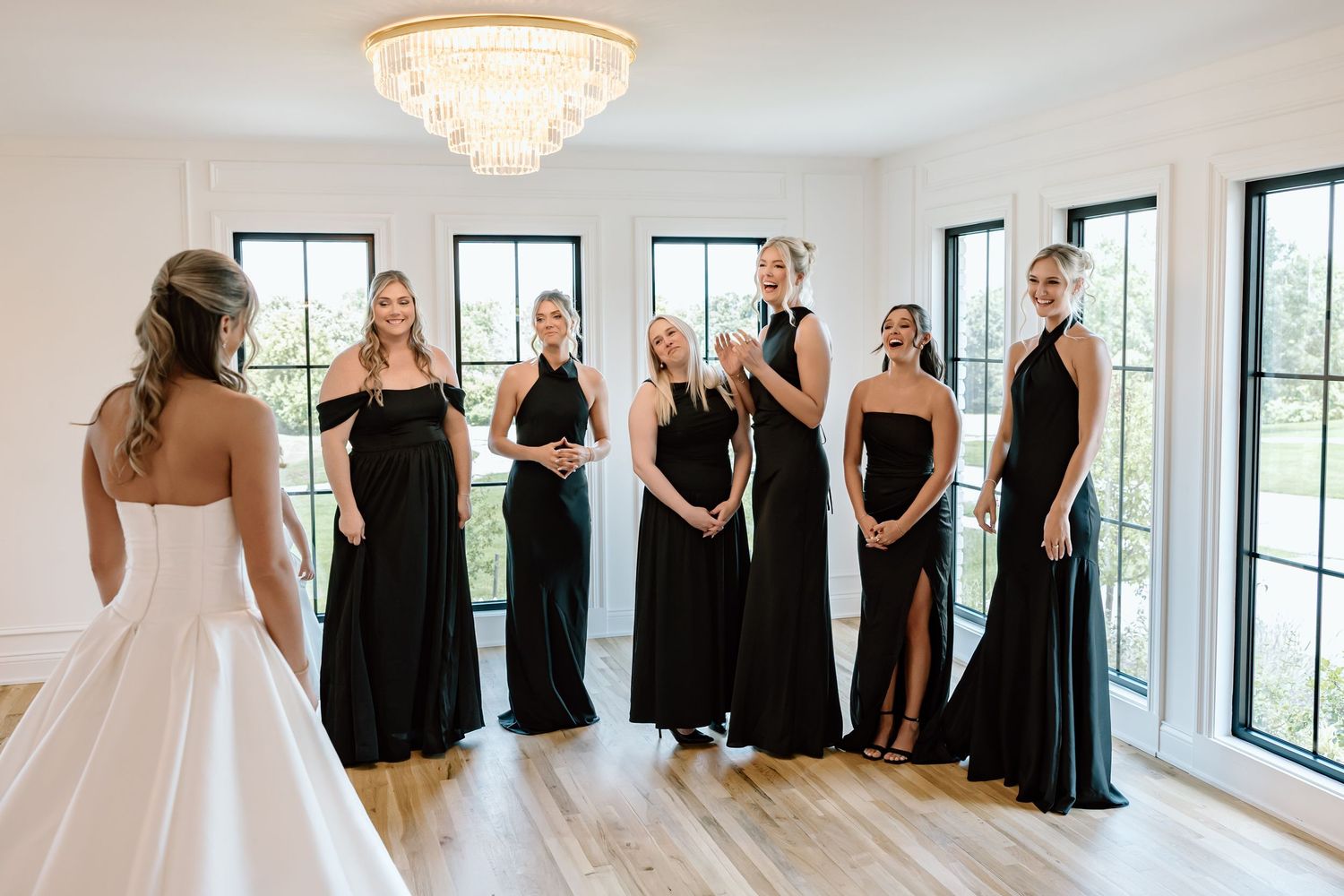 Bride in white gown reveals dress to bridesmaids in black dresses during first look in bright modern venue.