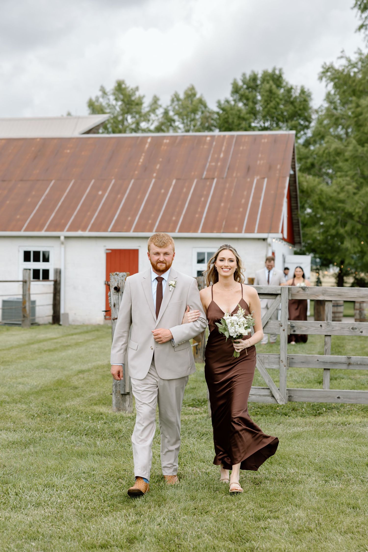 A groomsman in a tan suit escorts a bridesmaid in a brown dress across a green lawn at a rustic barn wedding.