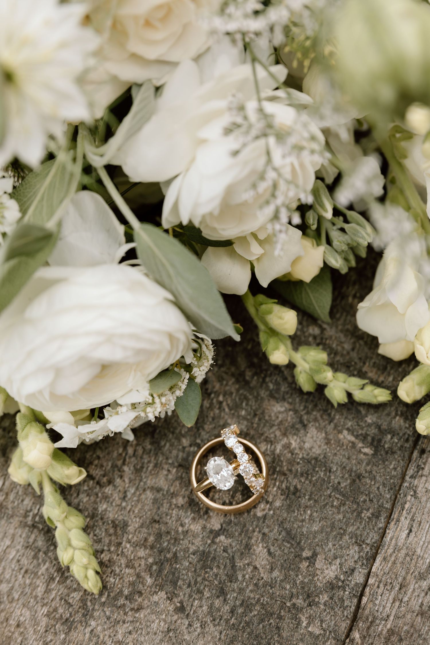 Gold wedding rings with diamond resting against white rose bridal bouquet on rustic wooden surface.