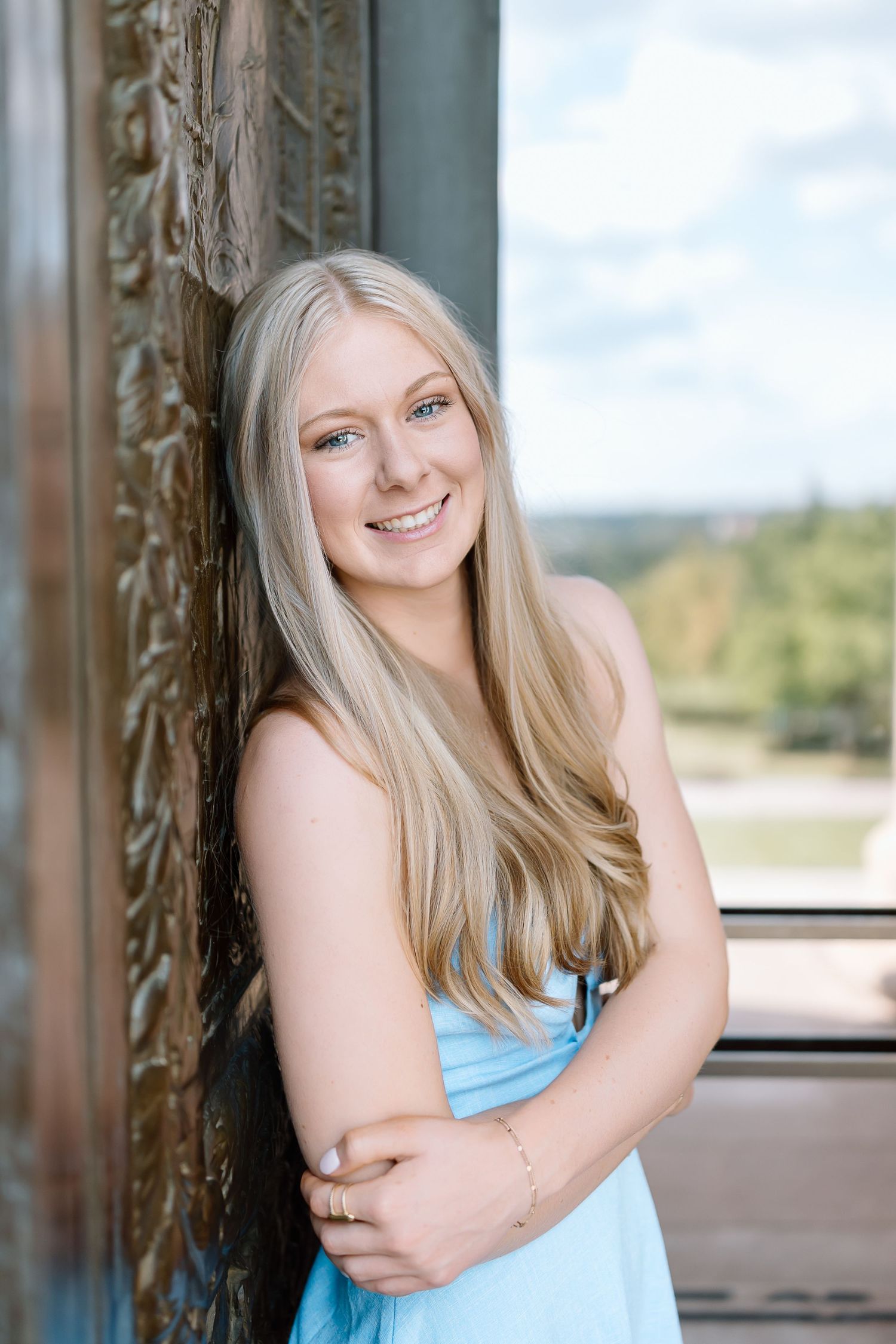 Portrait of someone with long blonde hair wearing a light blue dress standing by an ornate wooden doorframe.