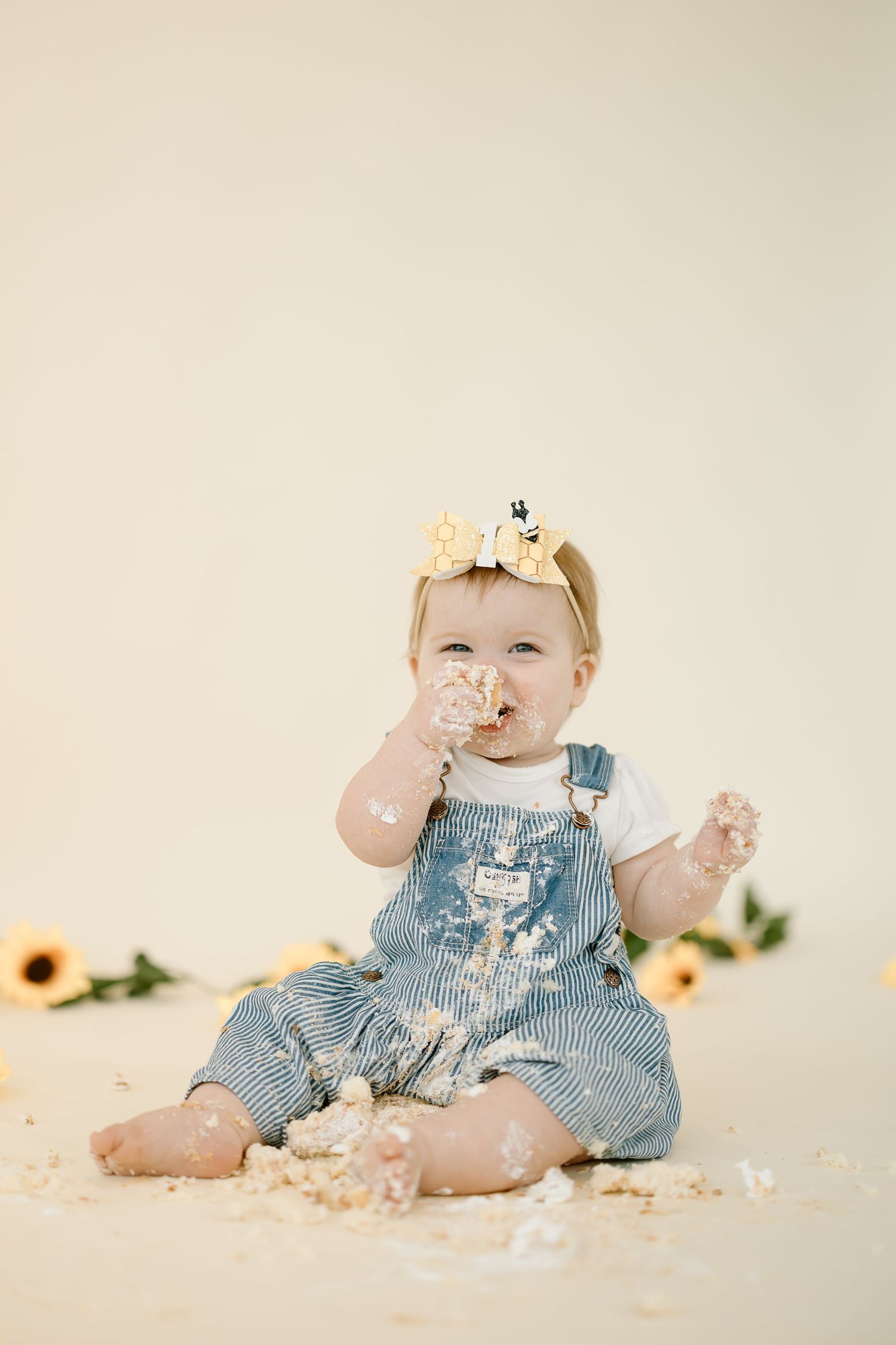 A baby in striped overalls sits on a cream backdrop while playing with cake smash during a first birthday photoshoot.