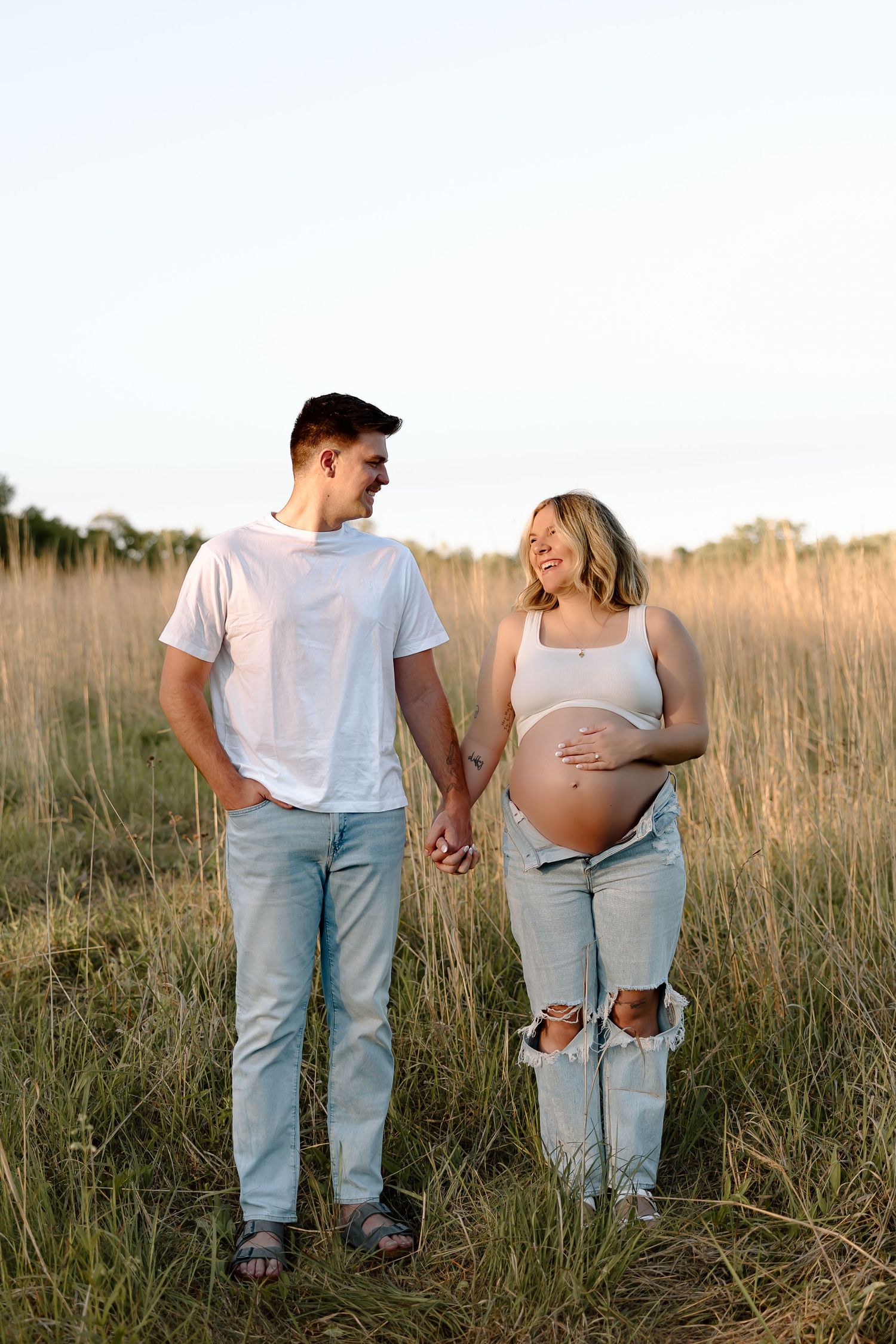Expecting couple holds hands while walking through tall grass field at sunset wearing casual denim and white outfits.