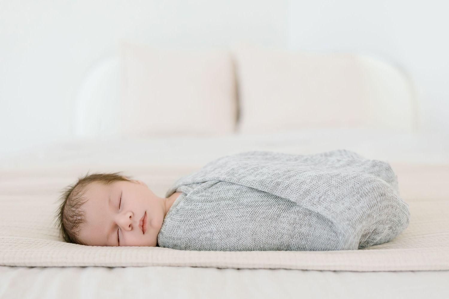 A sleeping baby wearing a gray sweater lies peacefully on white bedding.