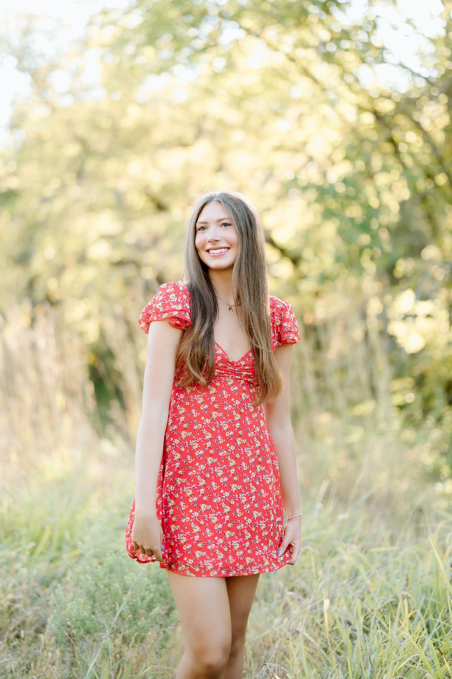 A person in a red floral dress poses outdoors in a sun-drenched field with tall grass and trees in the background.