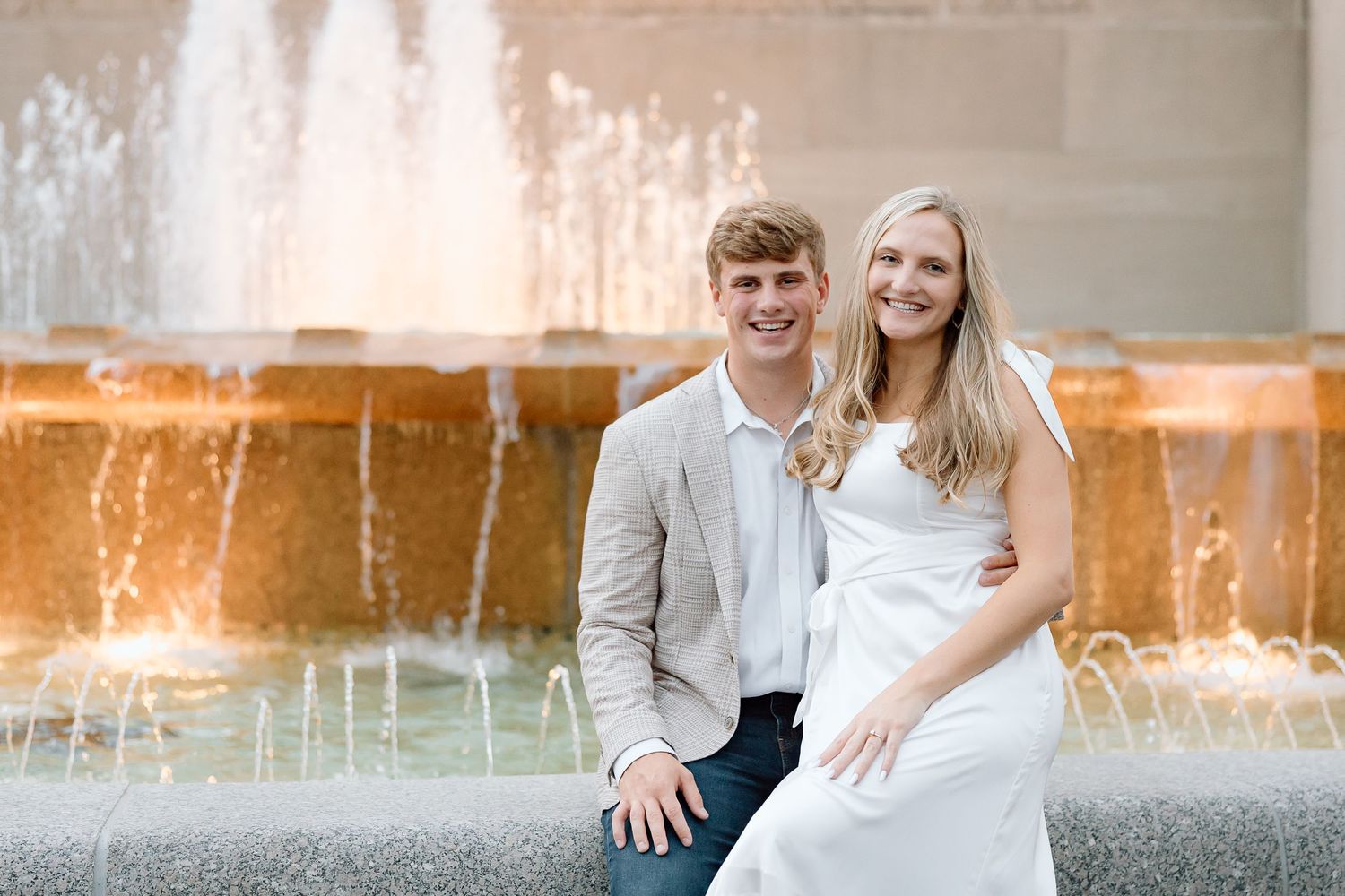 A couple poses for romantic engagement photos in front of a glowing fountain at sunset.