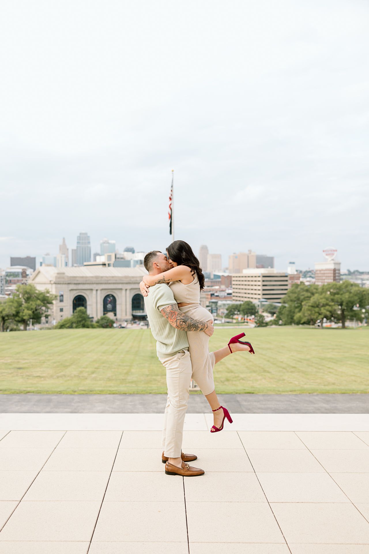 A romantic couple shares an embrace on a concrete plaza with a cityscape backdrop.