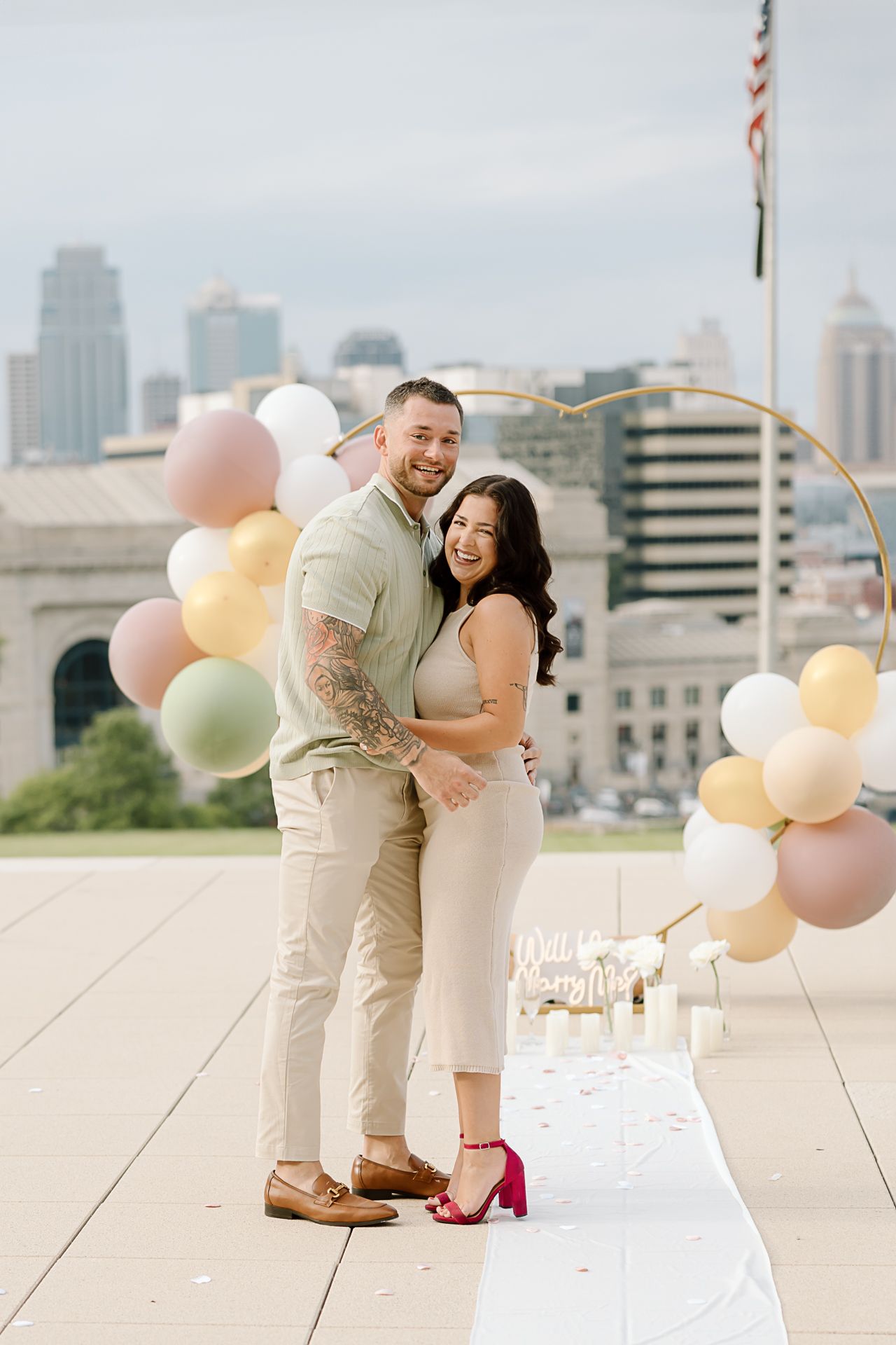 Sweet proposal moment captured on city rooftop decorated with pastel balloons and white runner.