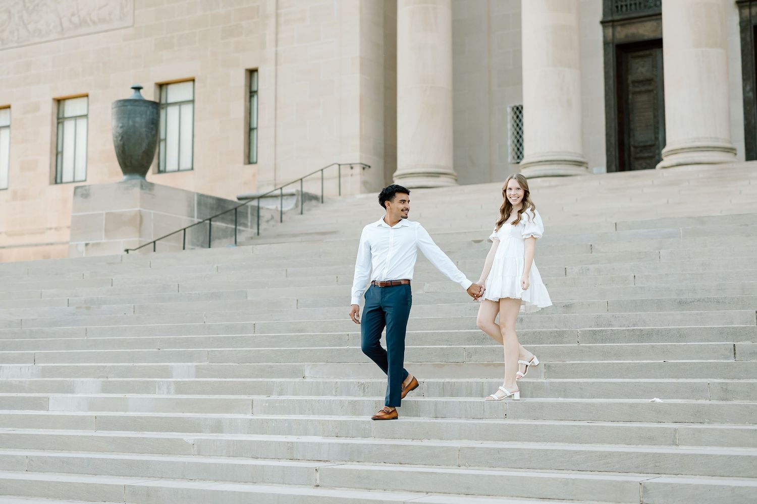 A couple in formal attire walks down stone steps outside a grand building with columns.