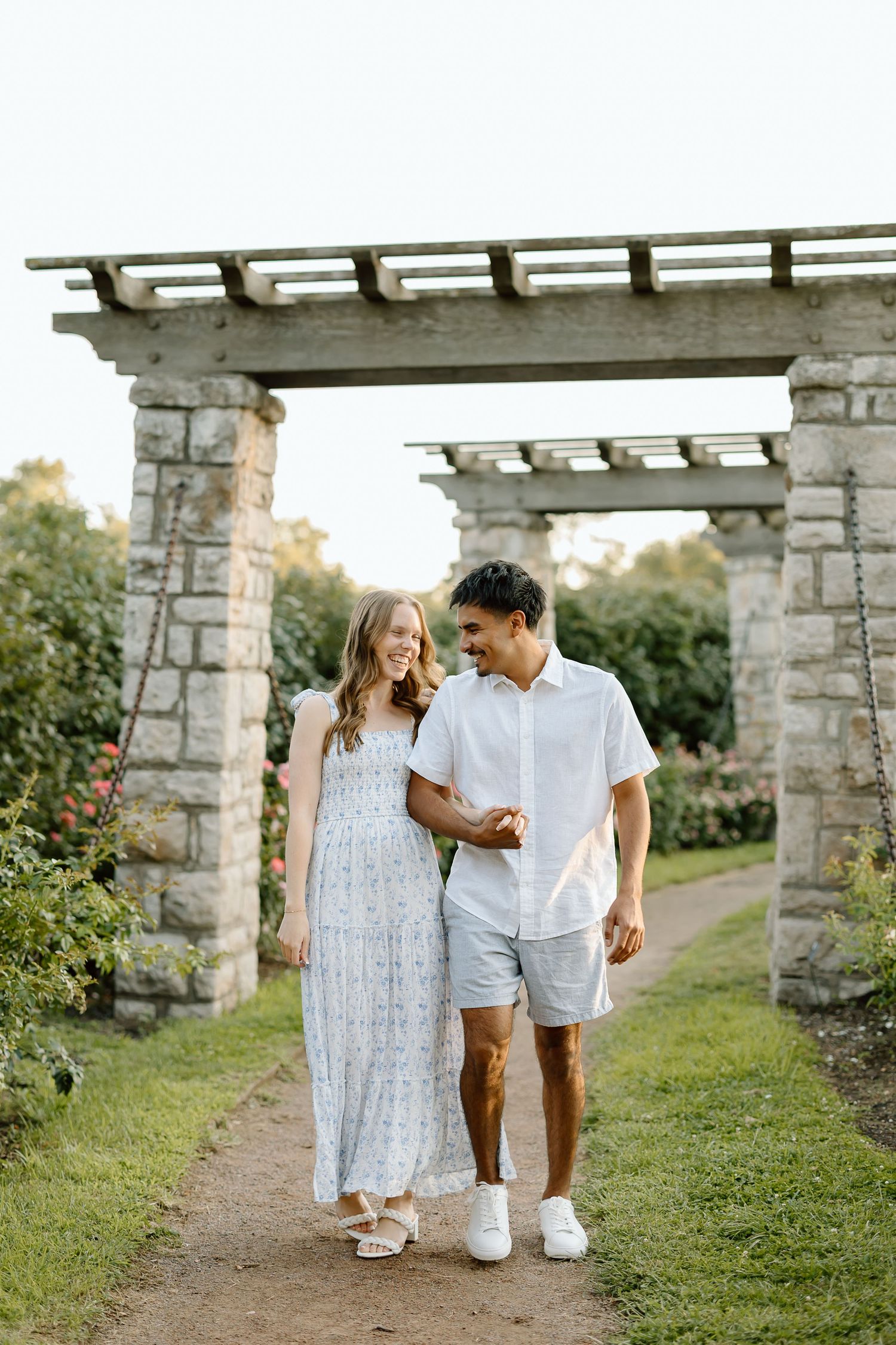 A couple walking together through a stone pergola in a garden setting wearing coordinated white and light blue outfits.