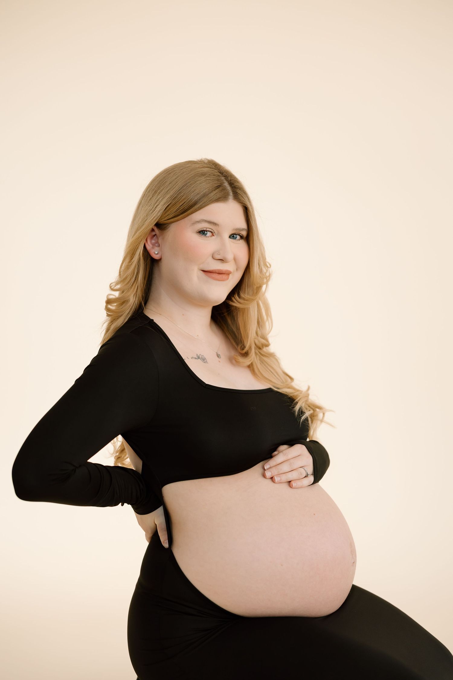 A pregnant woman in a black dress poses for a series of artistic maternity photos against a beige backdrop.
