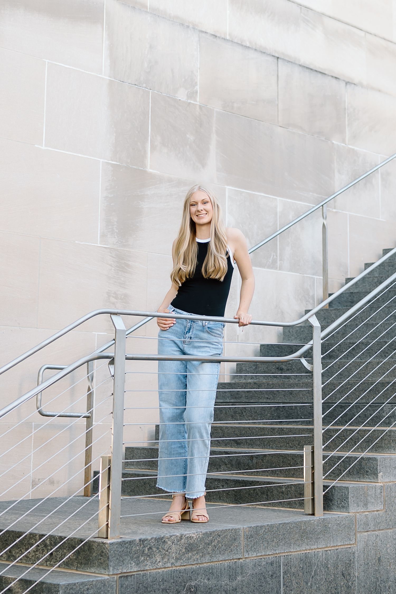 A person wearing jeans and a black top descends concrete stairs with metal railings in a minimalist setting.