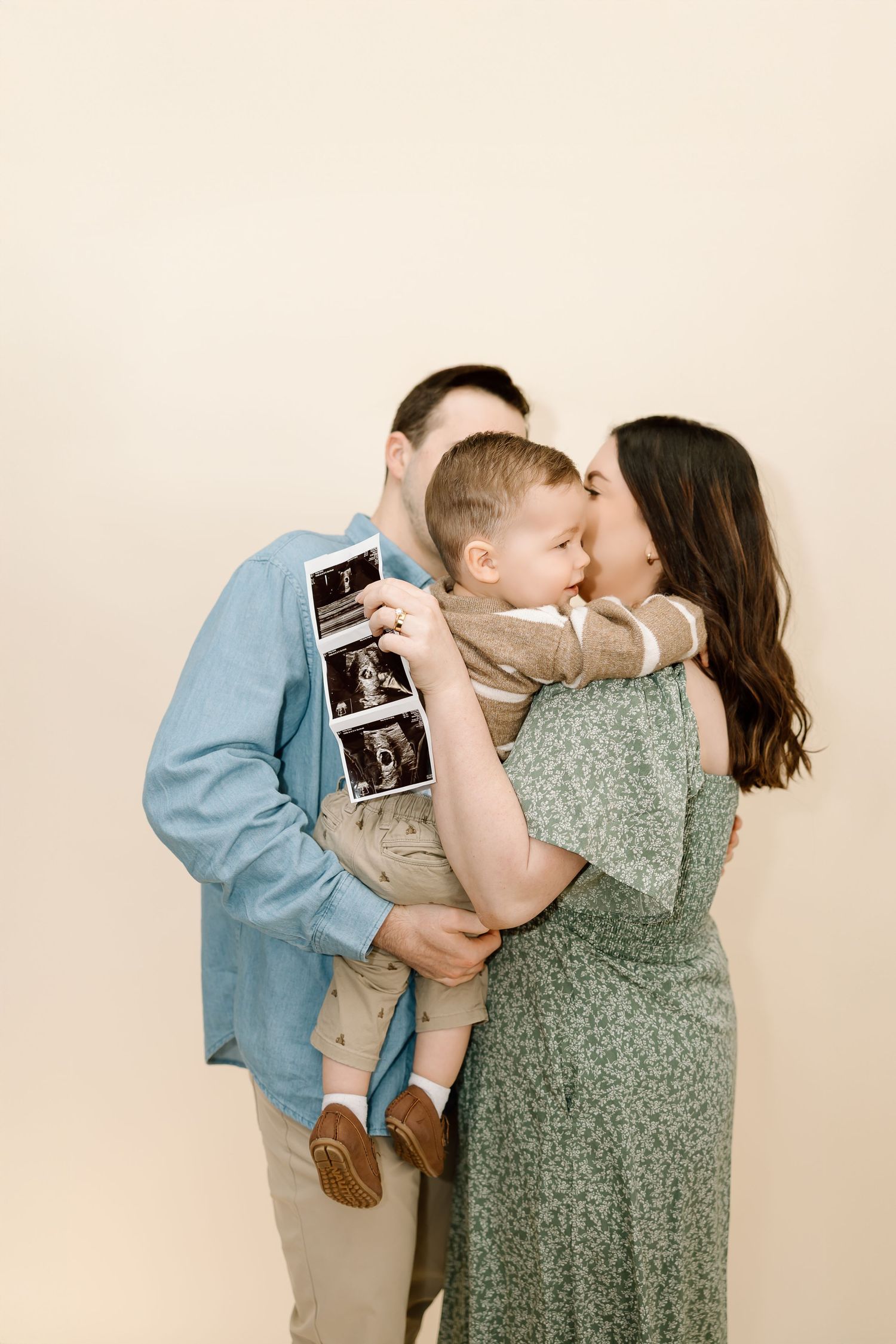 A couple in casual attire holds up ultrasound photos while sharing a tender moment in a studio setting.