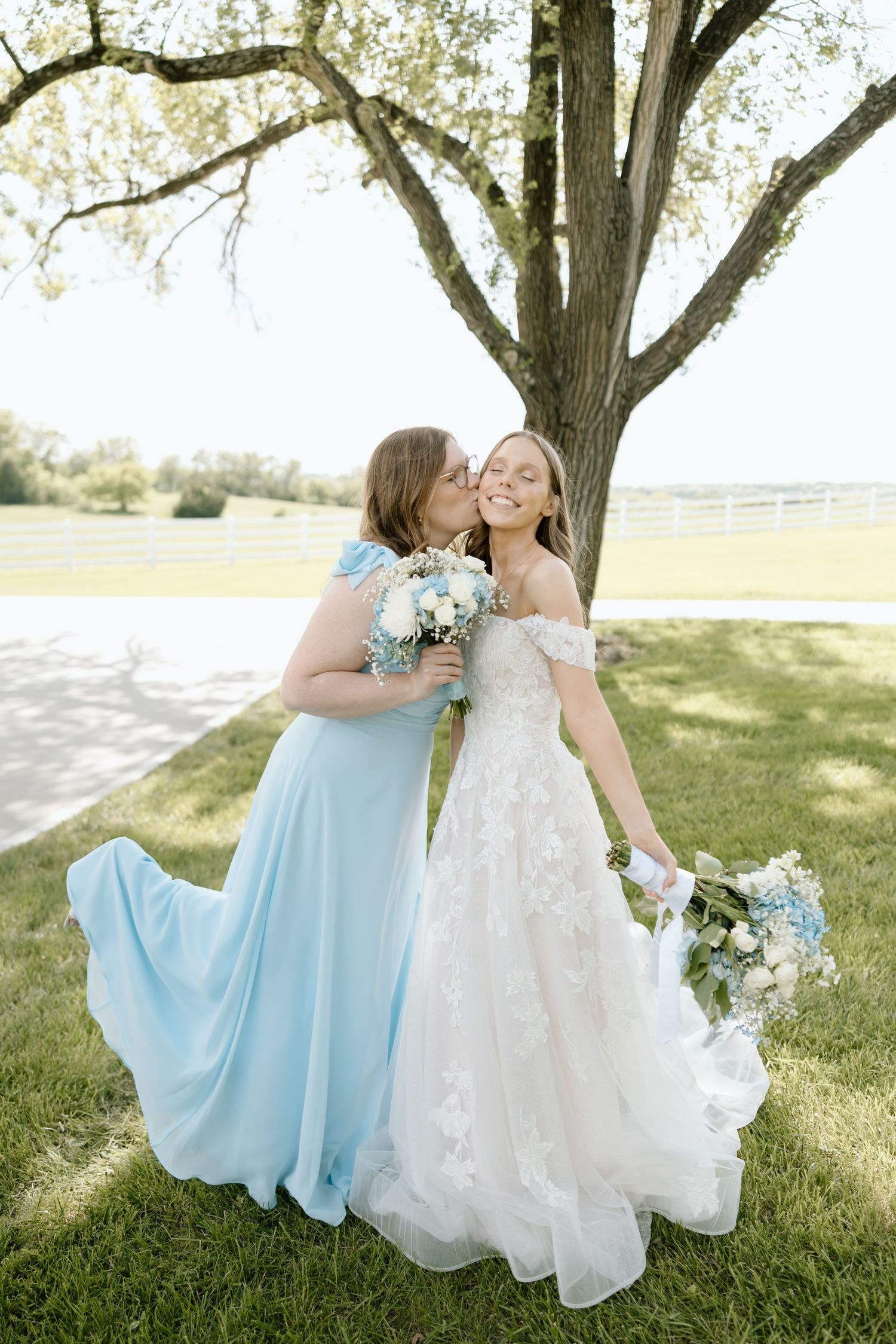 Bride in lace gown receives kiss on cheek from bridesmaid in light blue dress outdoors.