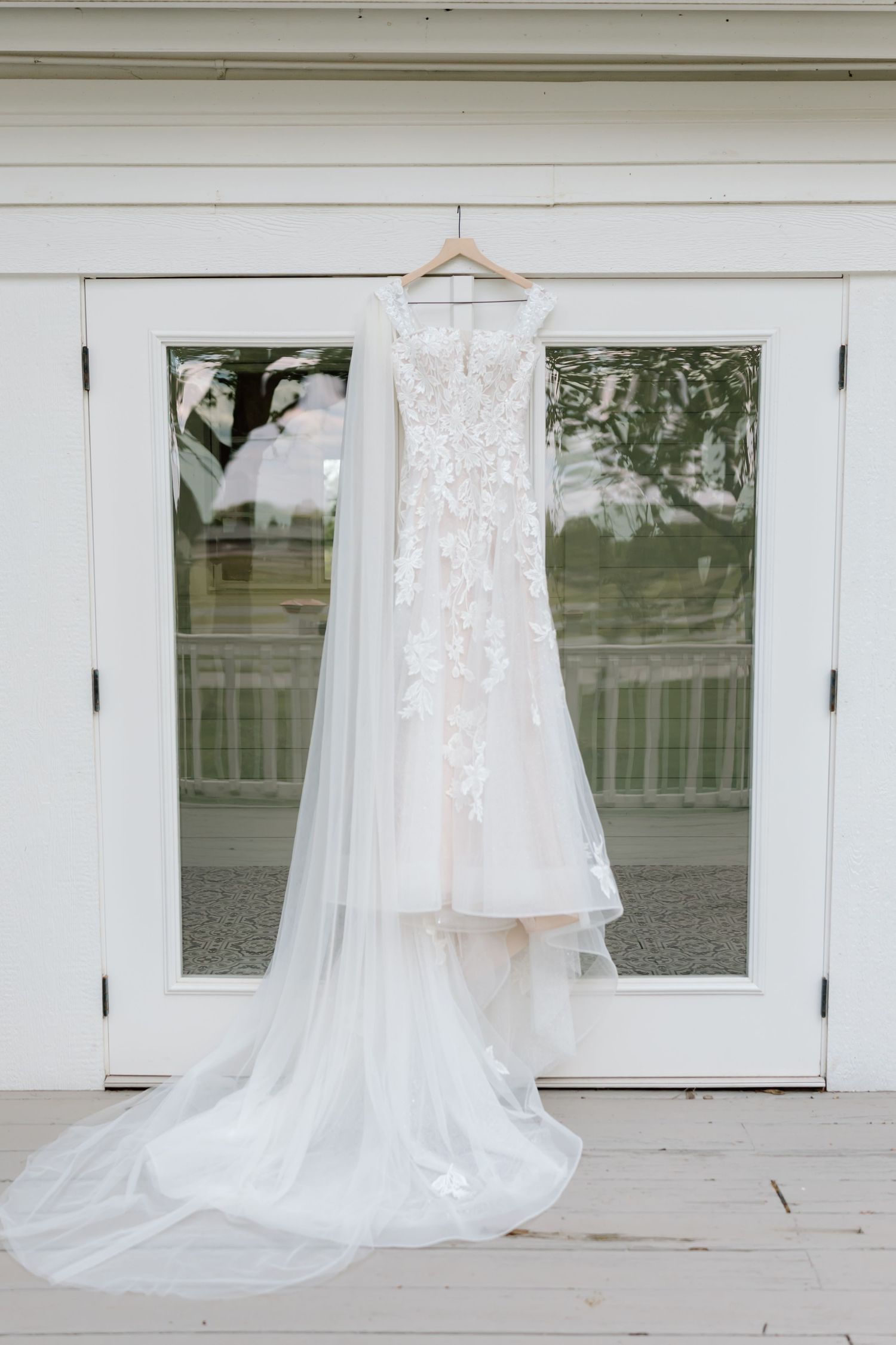 Elegant white lace wedding gown with long train hanging on a wooden hanger against glass French doors.