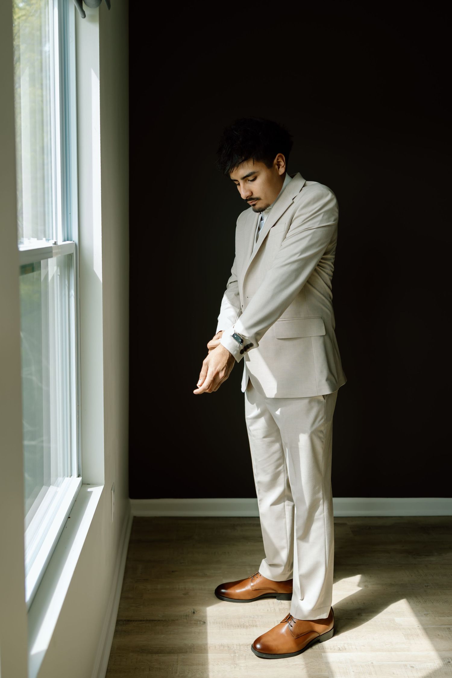 Man in white suit adjusting cufflinks by window in dark room, wearing brown leather dress shoes.