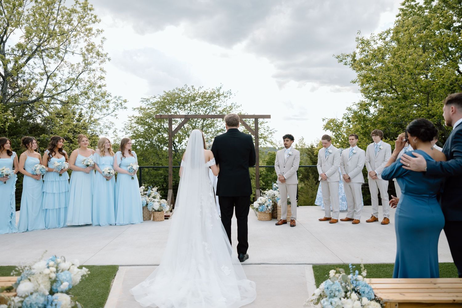 Outdoor wedding ceremony with bride in white gown, bridesmaids in blue dresses, and groomsmen in gray suits under cloudy sky.