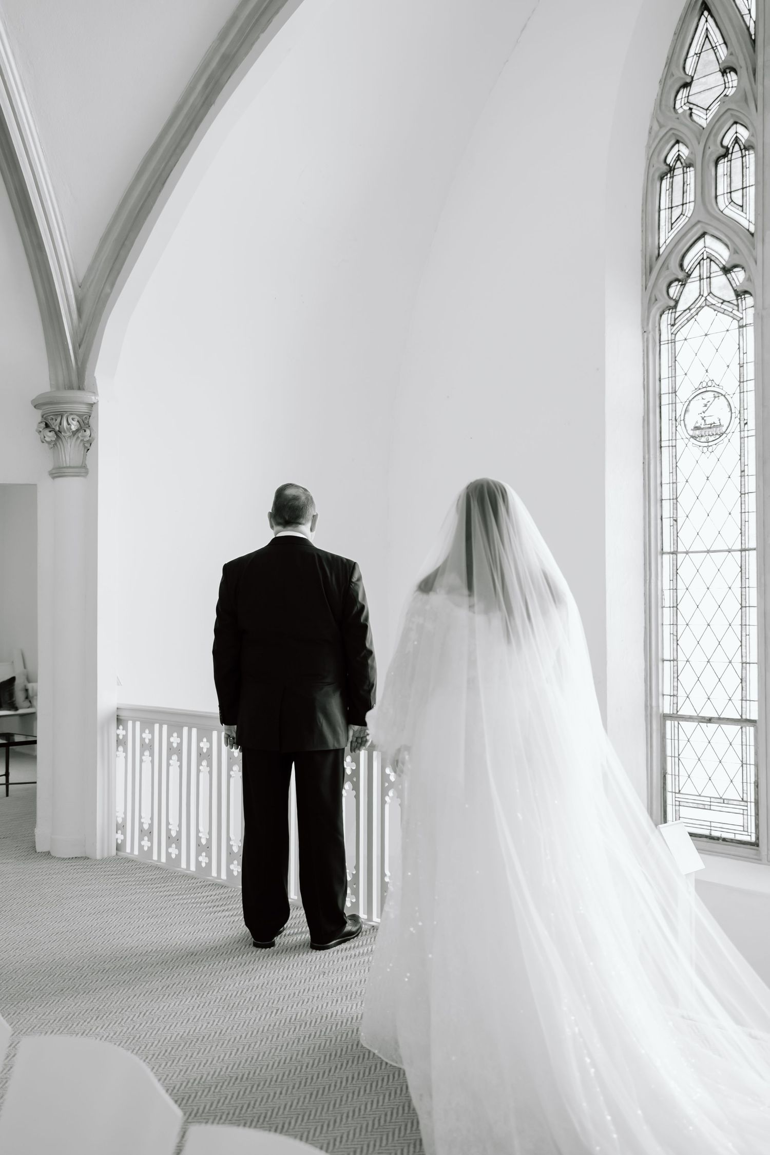 Black and white photo of a bride in a flowing veil and groom standing at church altar with stained glass windows.