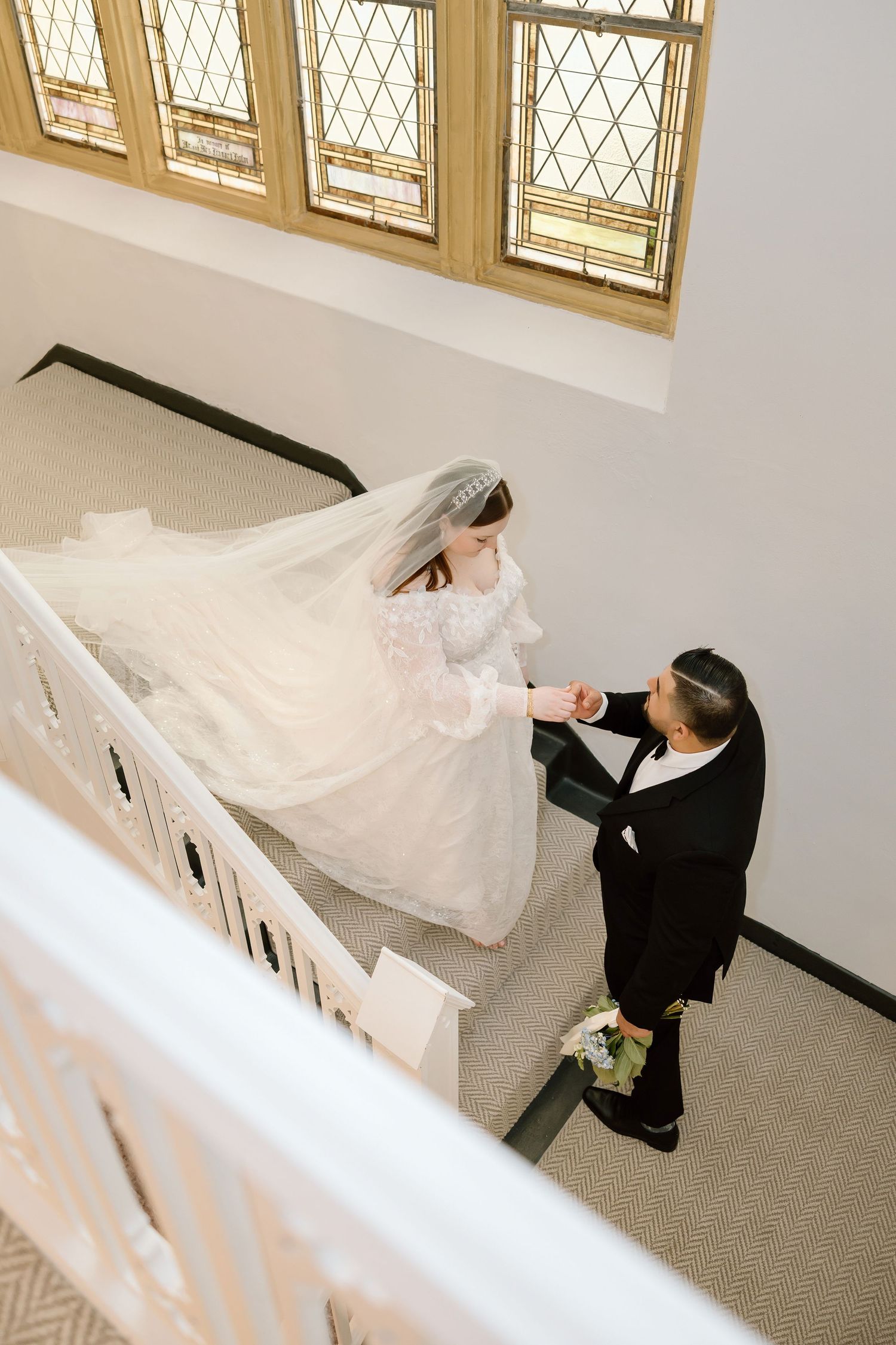 Bride in white gown and veil holds groom's hand on elegant staircase with stained glass windows above.