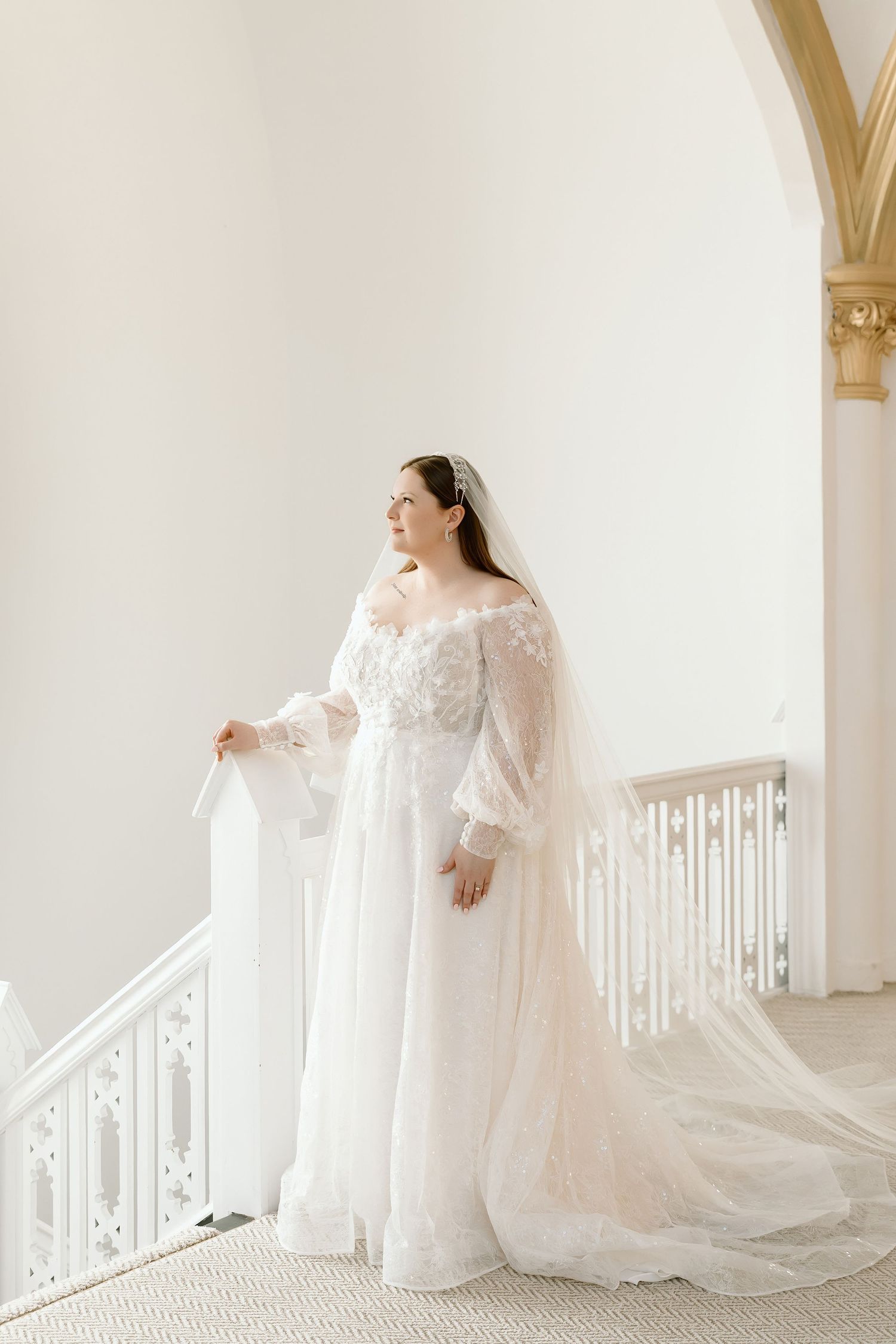 Bride in off-shoulder lace wedding gown with veil stands on elegant staircase in bright white venue.