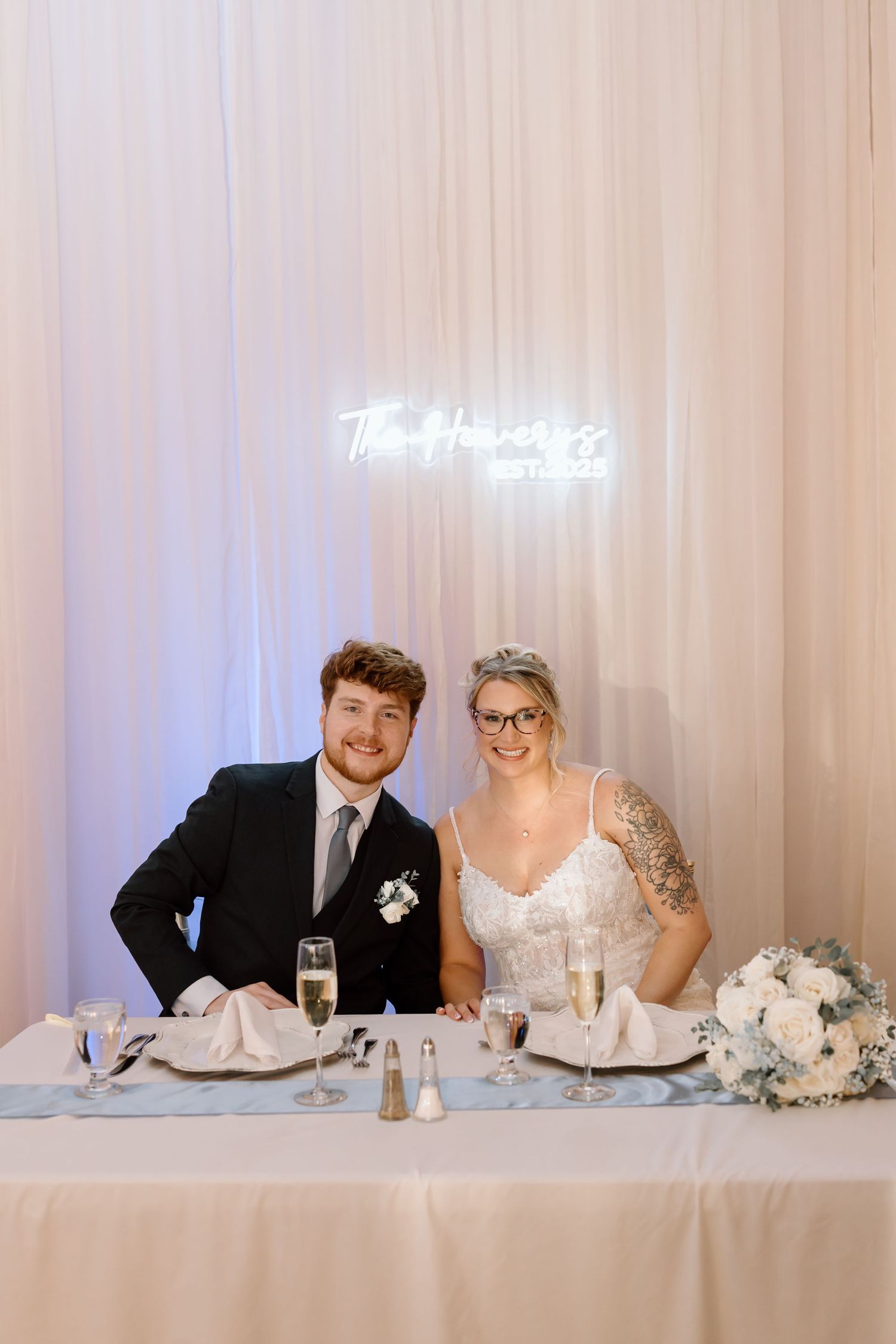 A couple sits at an elegant white-draped wedding reception head table with soft lighting and floral decor.