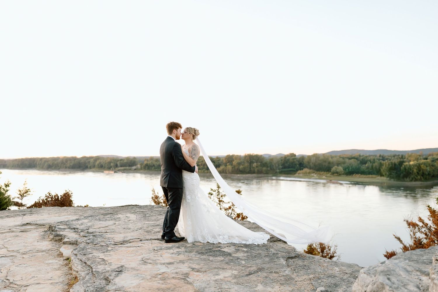 A couple in wedding attire embraces on a rocky shoreline with a serene lake and mountains in the background.
