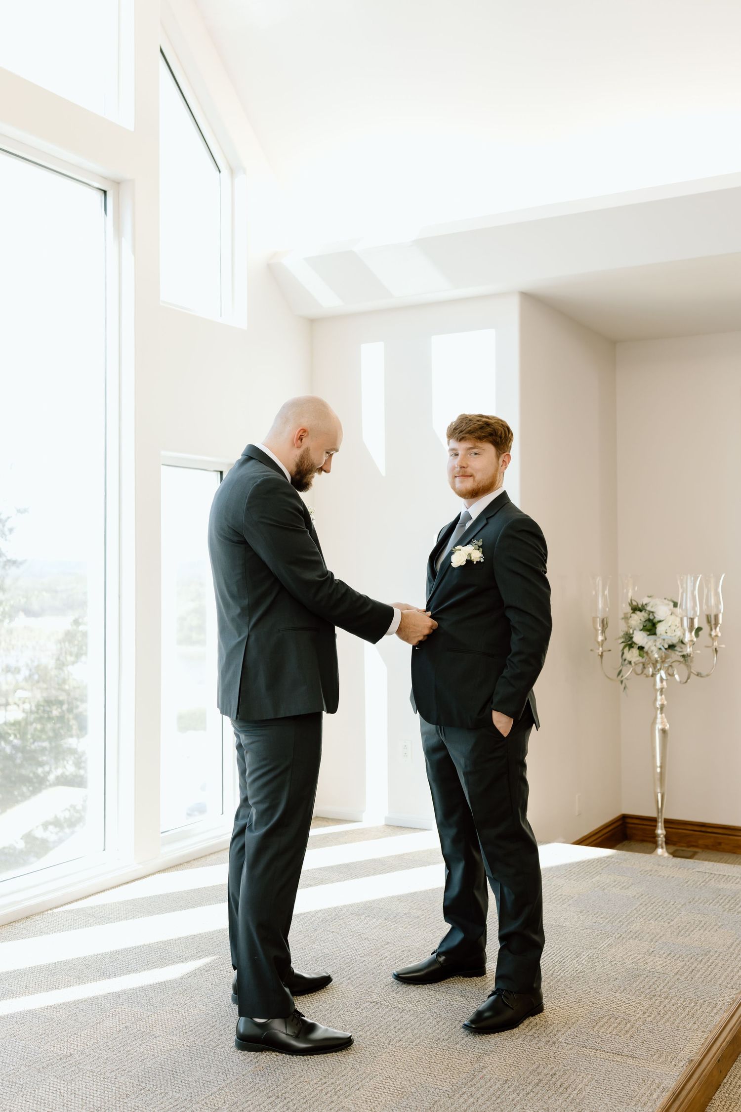 Two people in black suits adjust each other's ties in a bright white room with large windows.