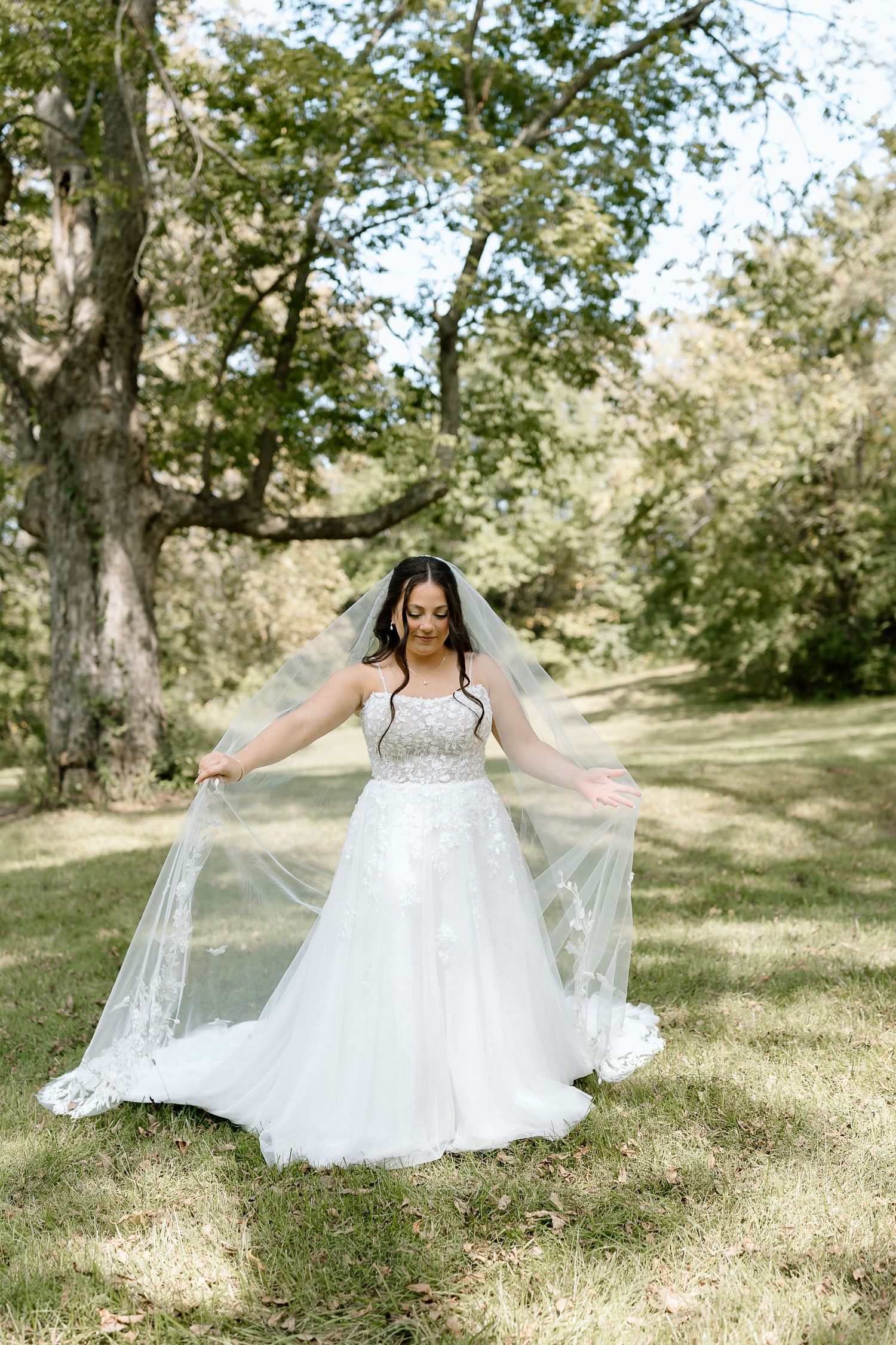 A bride in a flowing white wedding gown with a long veil poses gracefully outdoors among trees in a sunlit garden setting.
