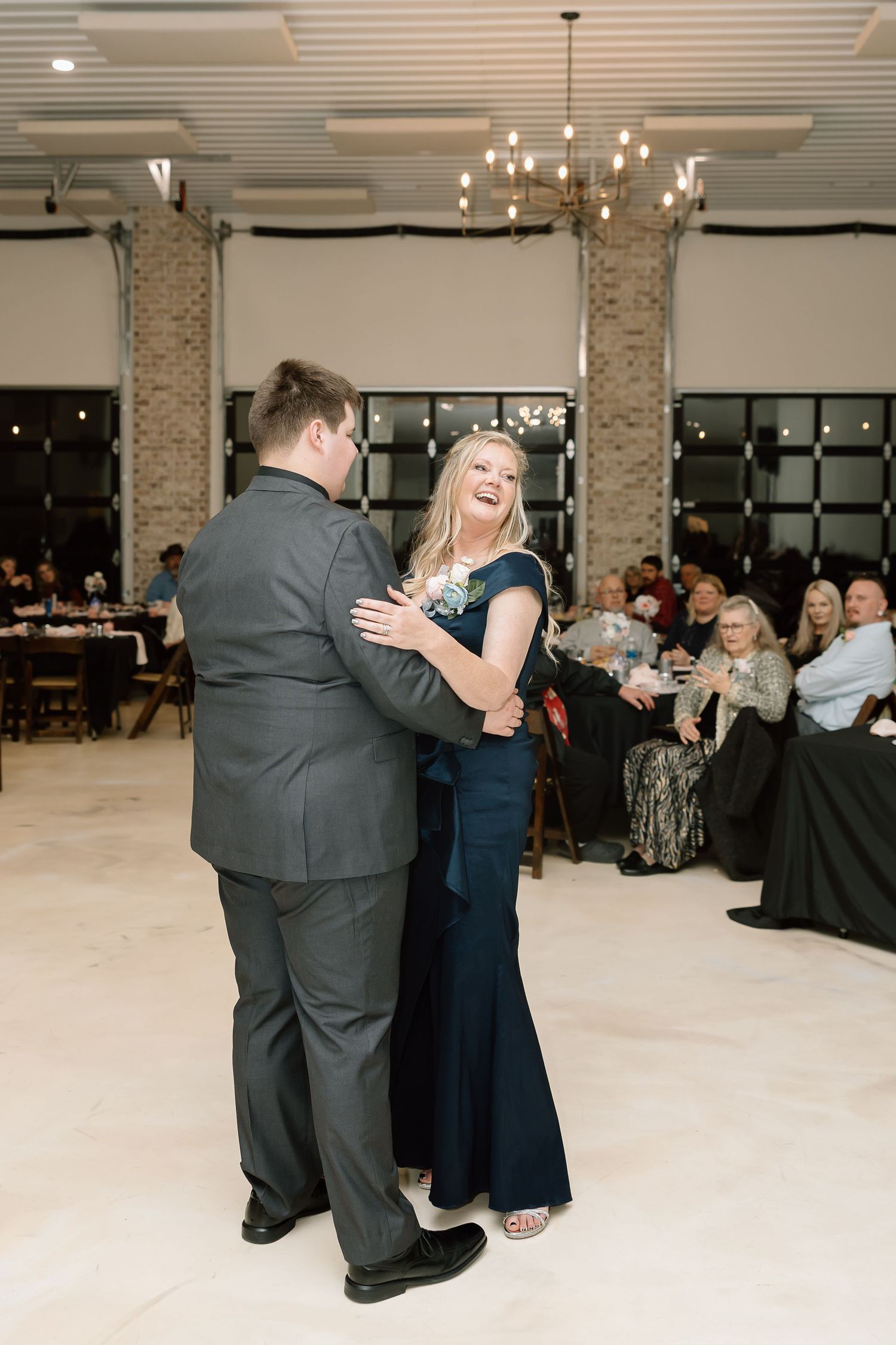 A couple dances together at a formal event while guests watch from tables in an elegant venue with exposed brick walls.