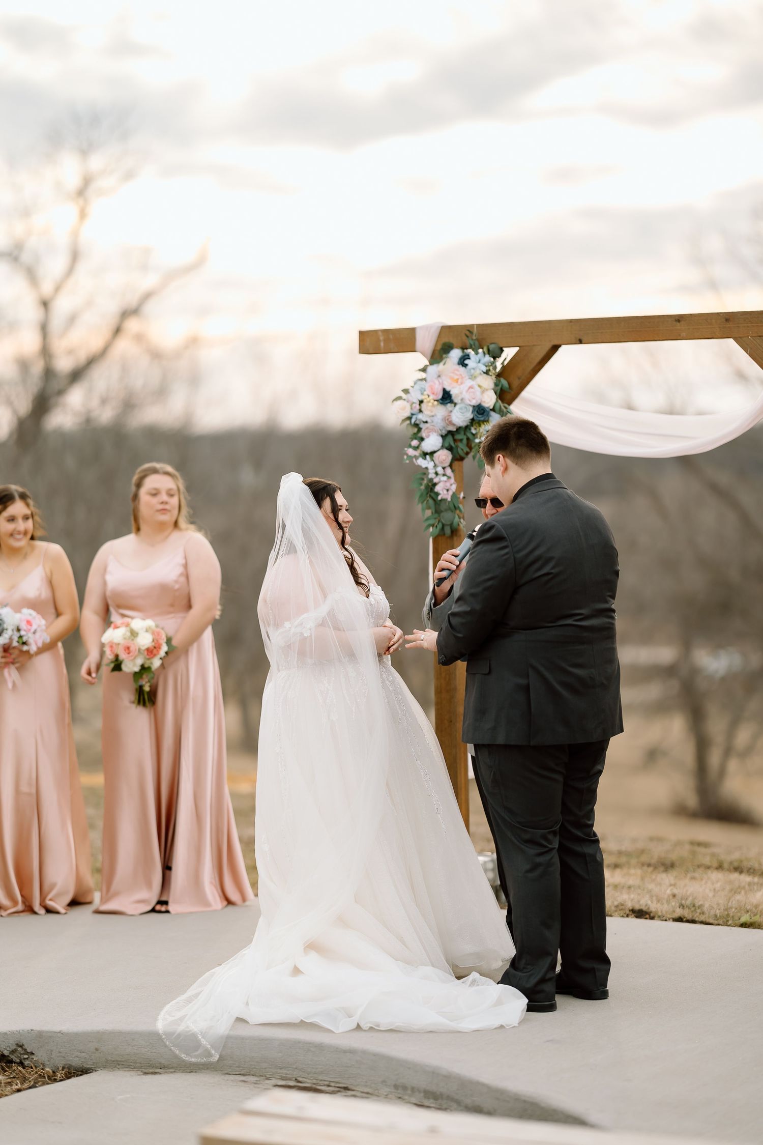 A wedding ceremony with bridesmaids in blush pink dresses watching the couple exchange vows under a wooden arch outdoors.