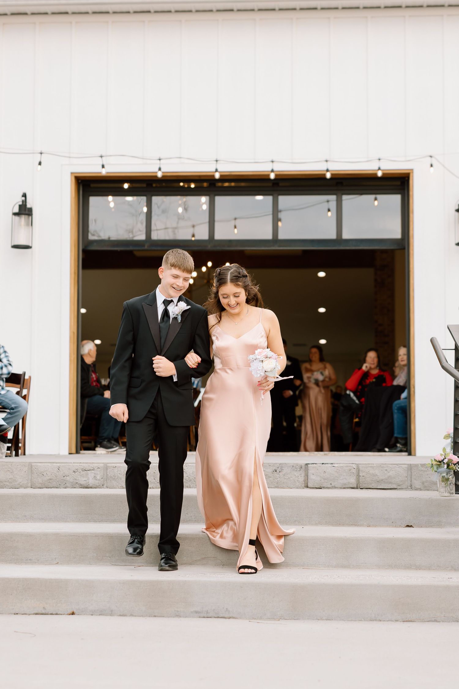 A couple walks together in formal attire outside a modern building with glass doors and windows.
