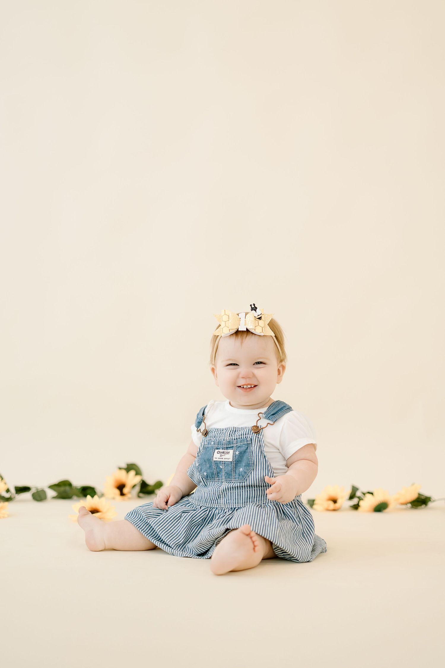 Small child wearing light blue denim overalls and white shirt sits on cream backdrop with floral decorations.