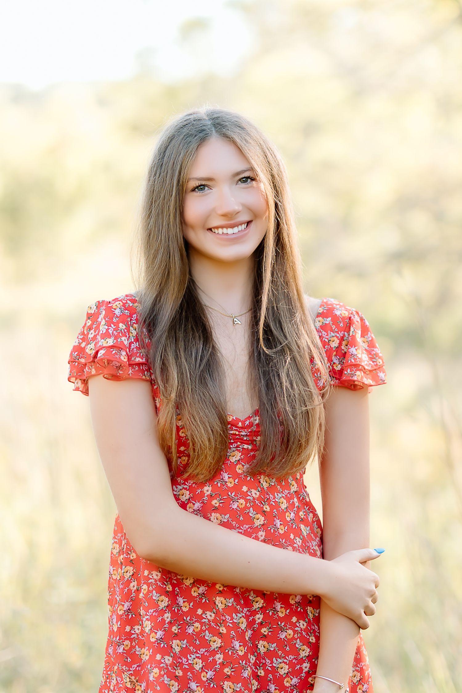 A person wearing a red floral dress poses outdoors in a sunny field with a natural bokeh background.