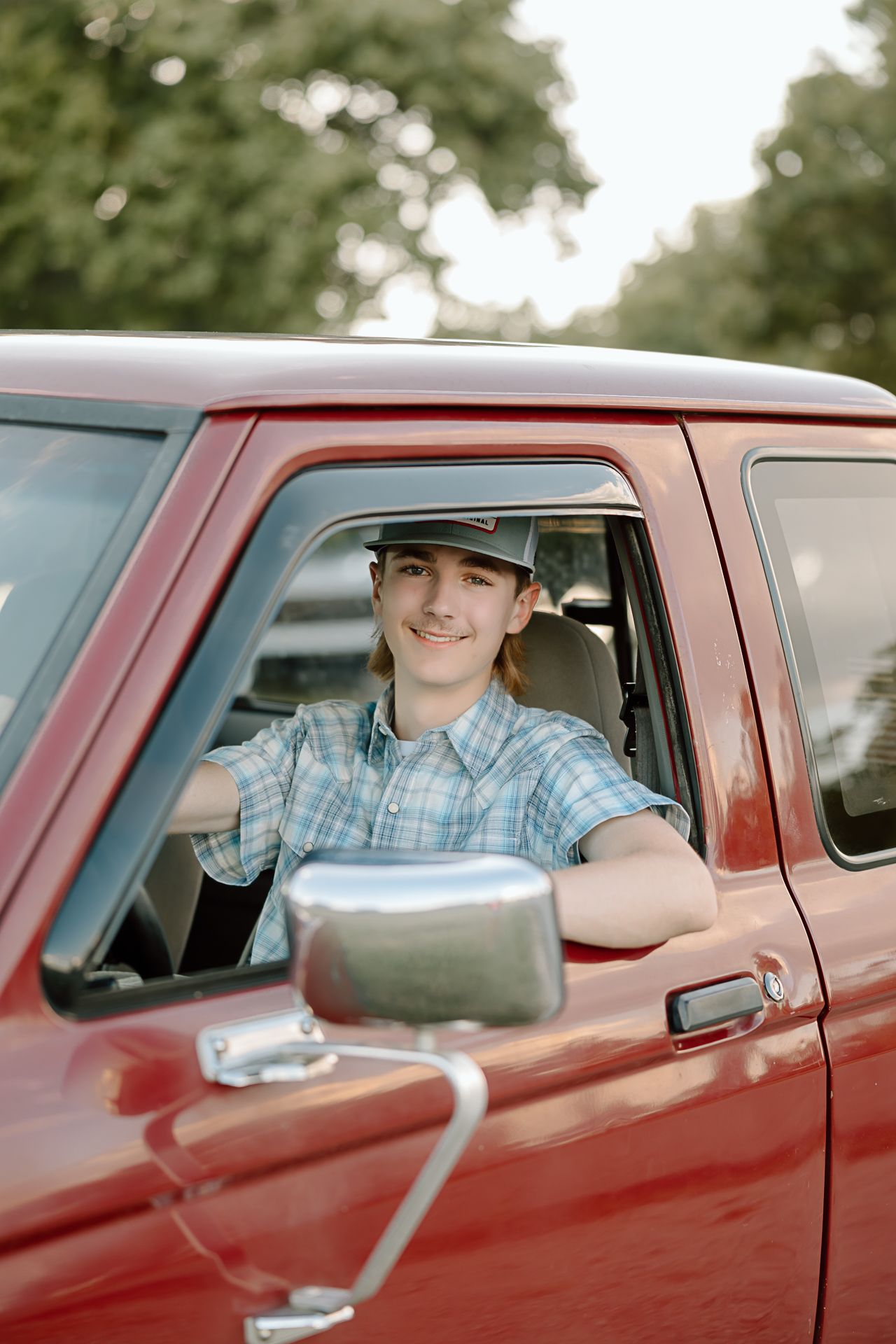 Someone sits in the driver's seat of a vintage red pickup truck wearing a plaid shirt and cowboy hat.