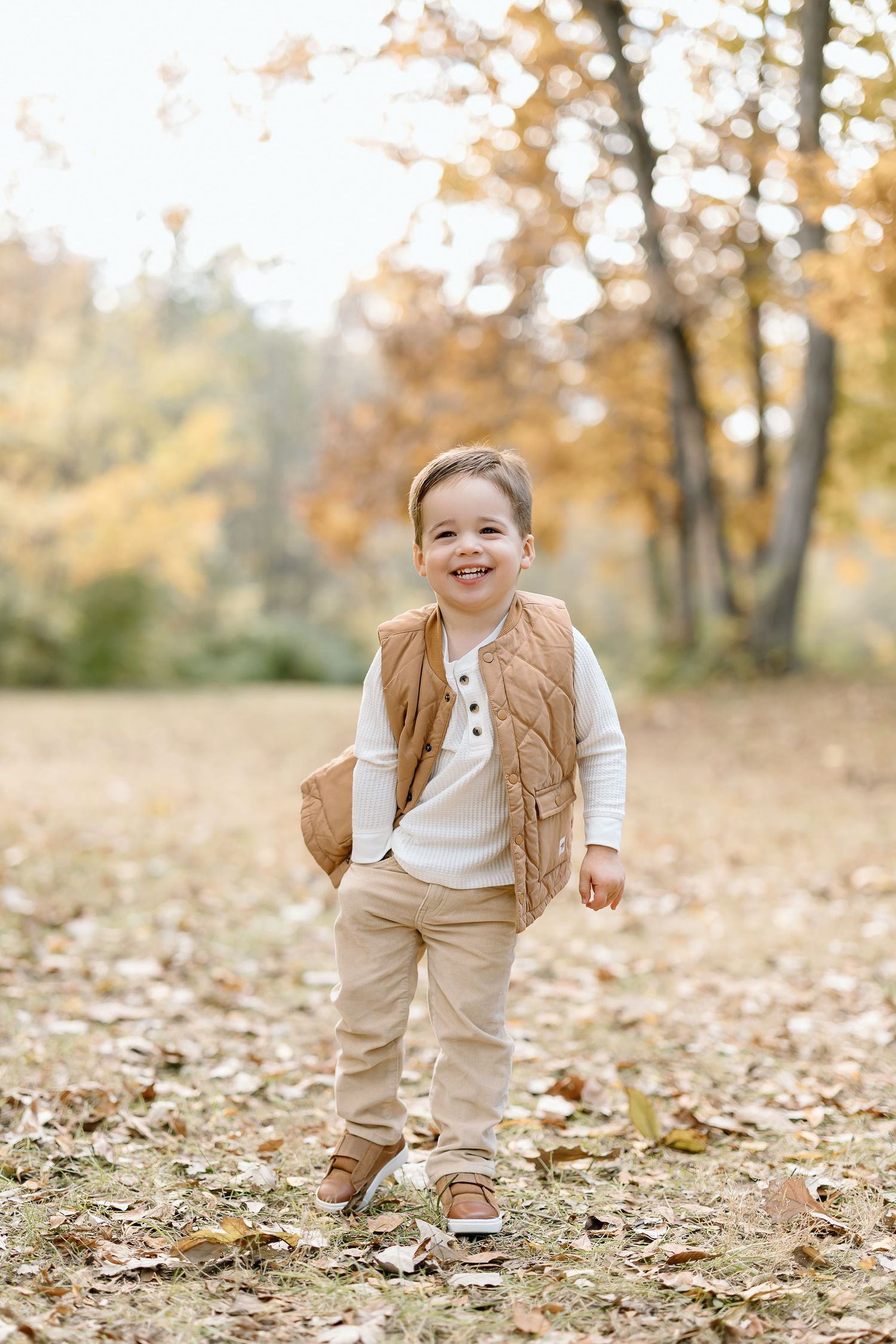 A child in khaki overalls and white shirt plays joyfully among fallen autumn leaves in a wooded setting.