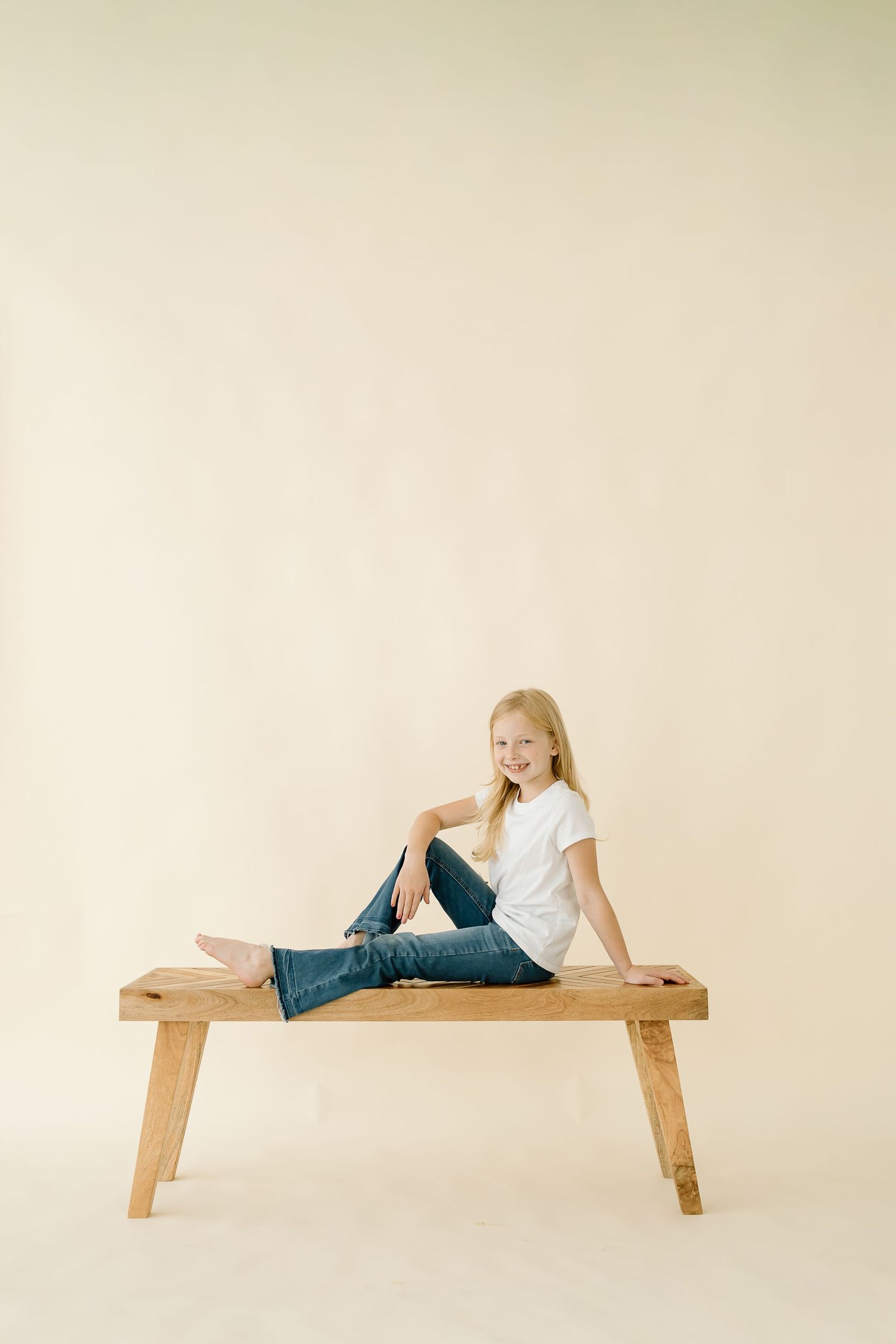 Child in white shirt and jeans sits casually on wooden bench against neutral beige background.
