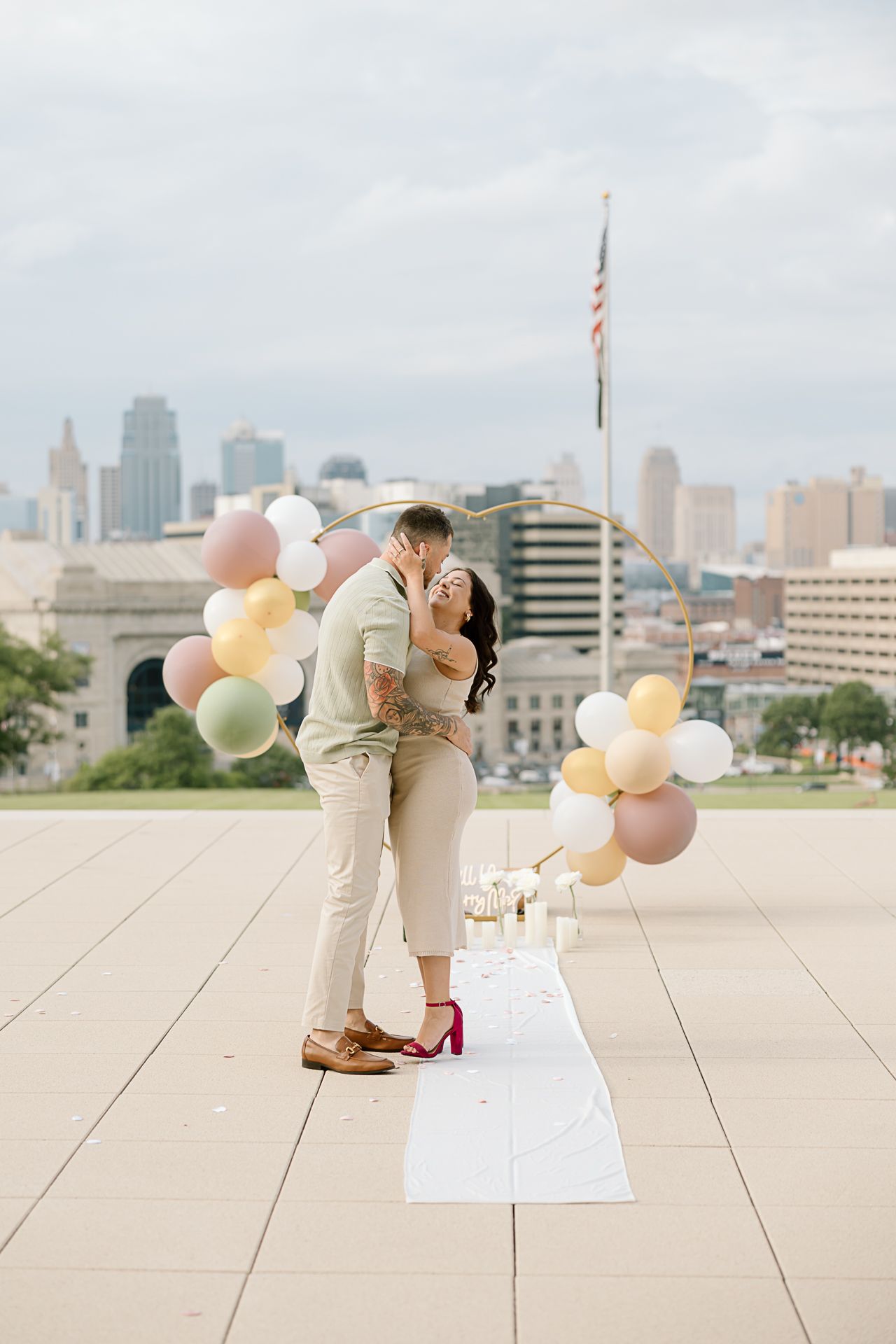 Couple sharing a romantic moment on urban rooftop with pastel balloon decorations and city views.