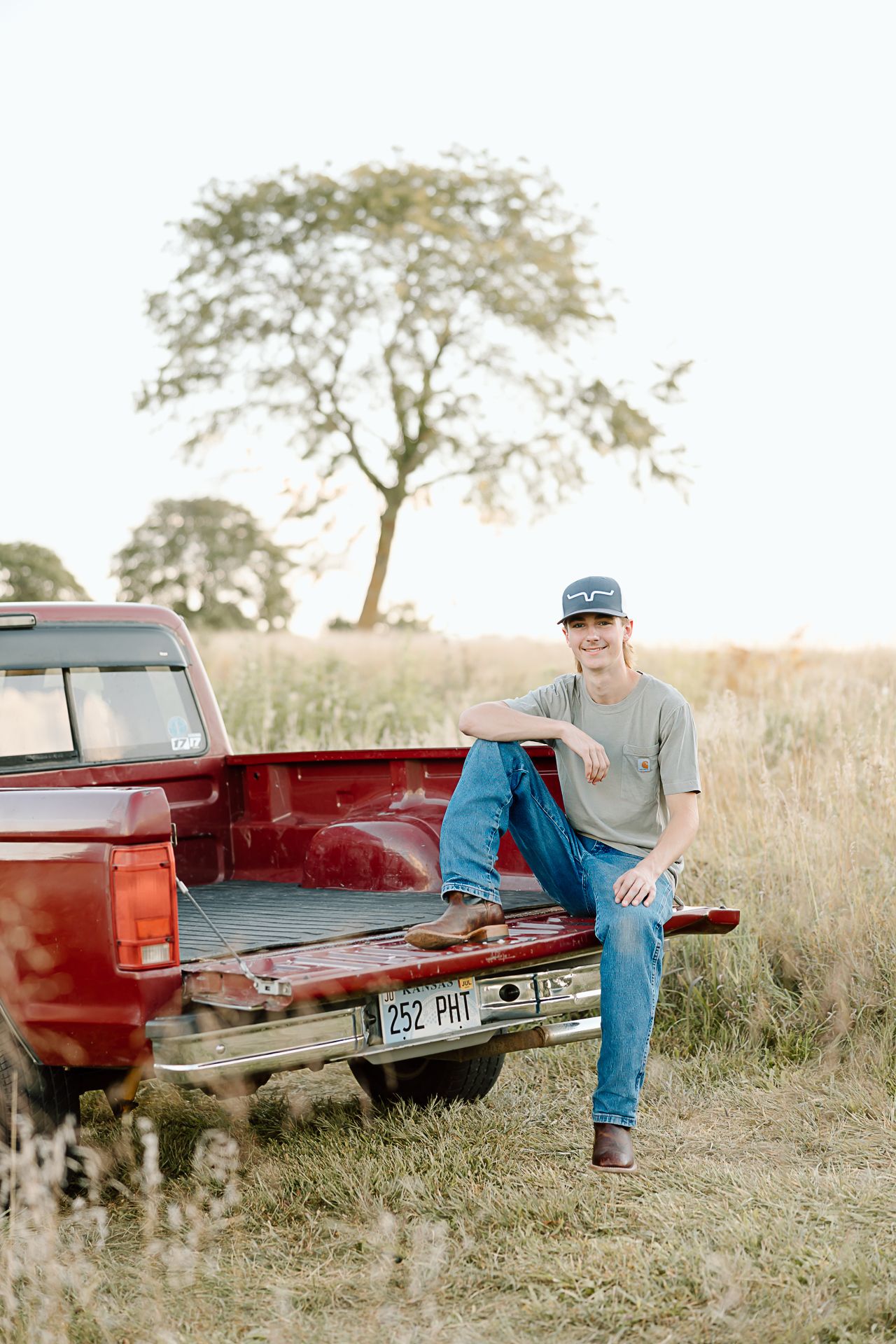 A person sits casually in the bed of a vintage red pickup truck parked in a rural field with a tree in the background.