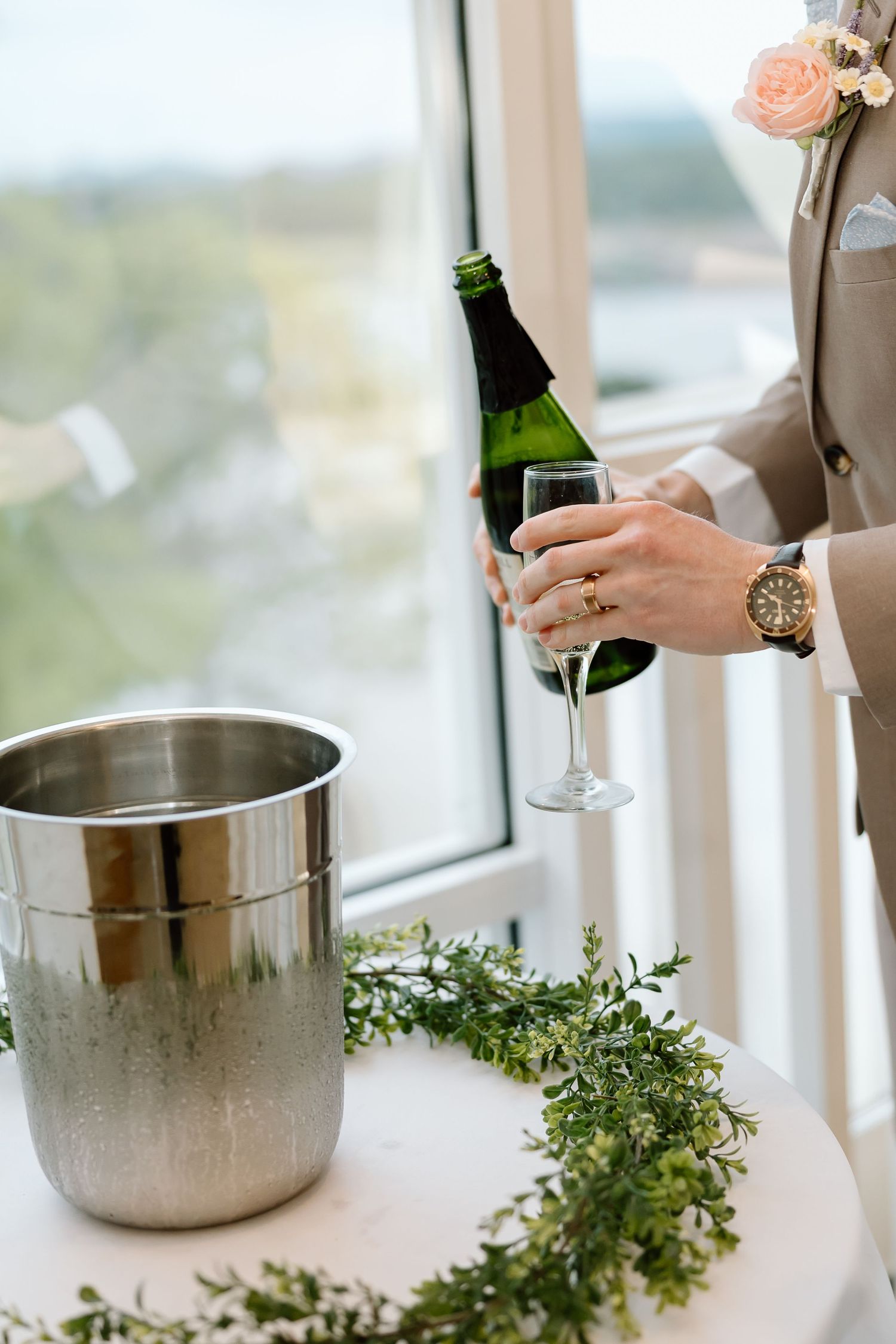 A hand holds a champagne bottle near an ice bucket with fresh herbs and glassware on a white table.