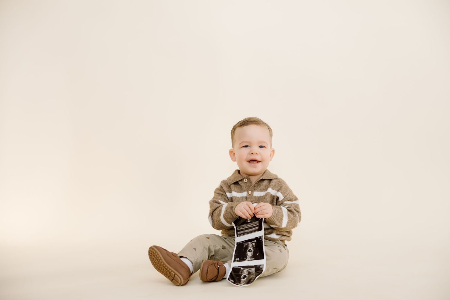 A smiling baby in a beige romper sits on the floor holding an ultrasound picture against a light background.