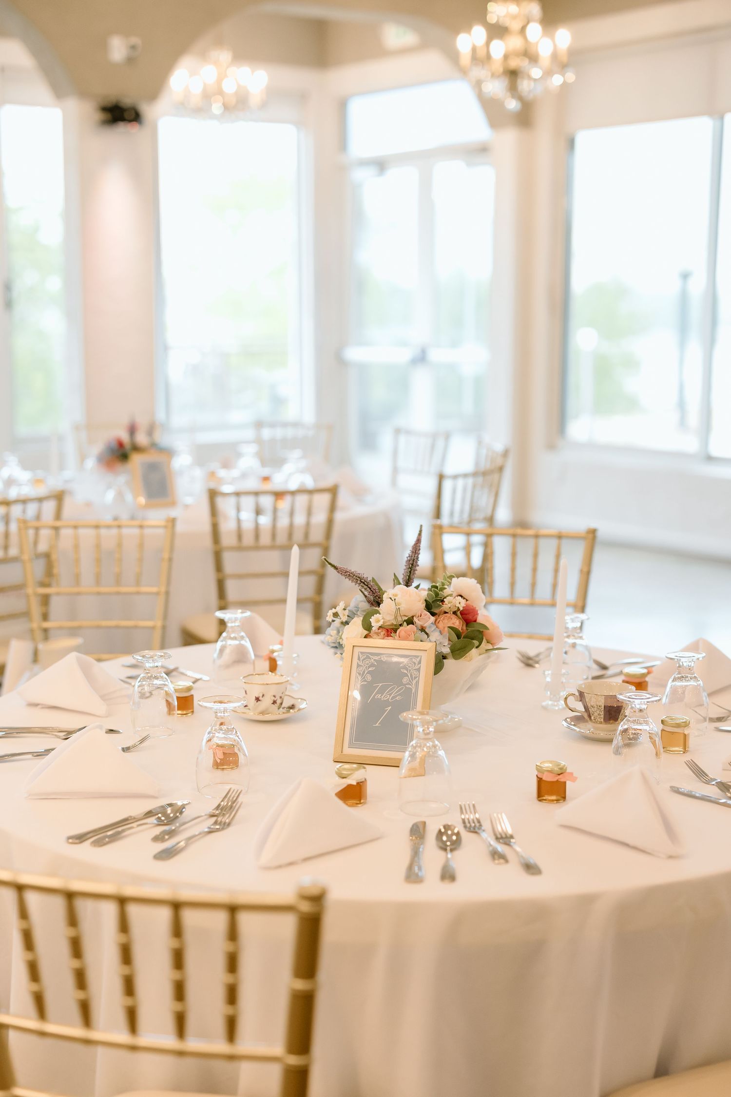 Elegant white reception tables with gold chiavari chairs and place settings in a bright banquet hall with chandeliers.