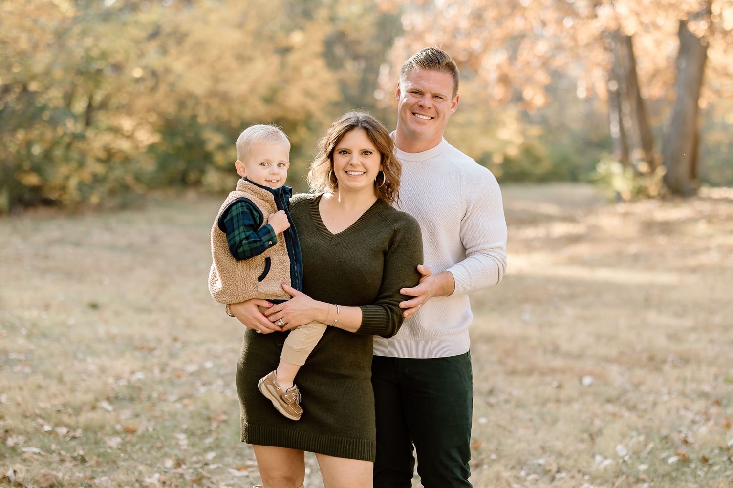 Family of three poses together in casual attire against autumn trees.