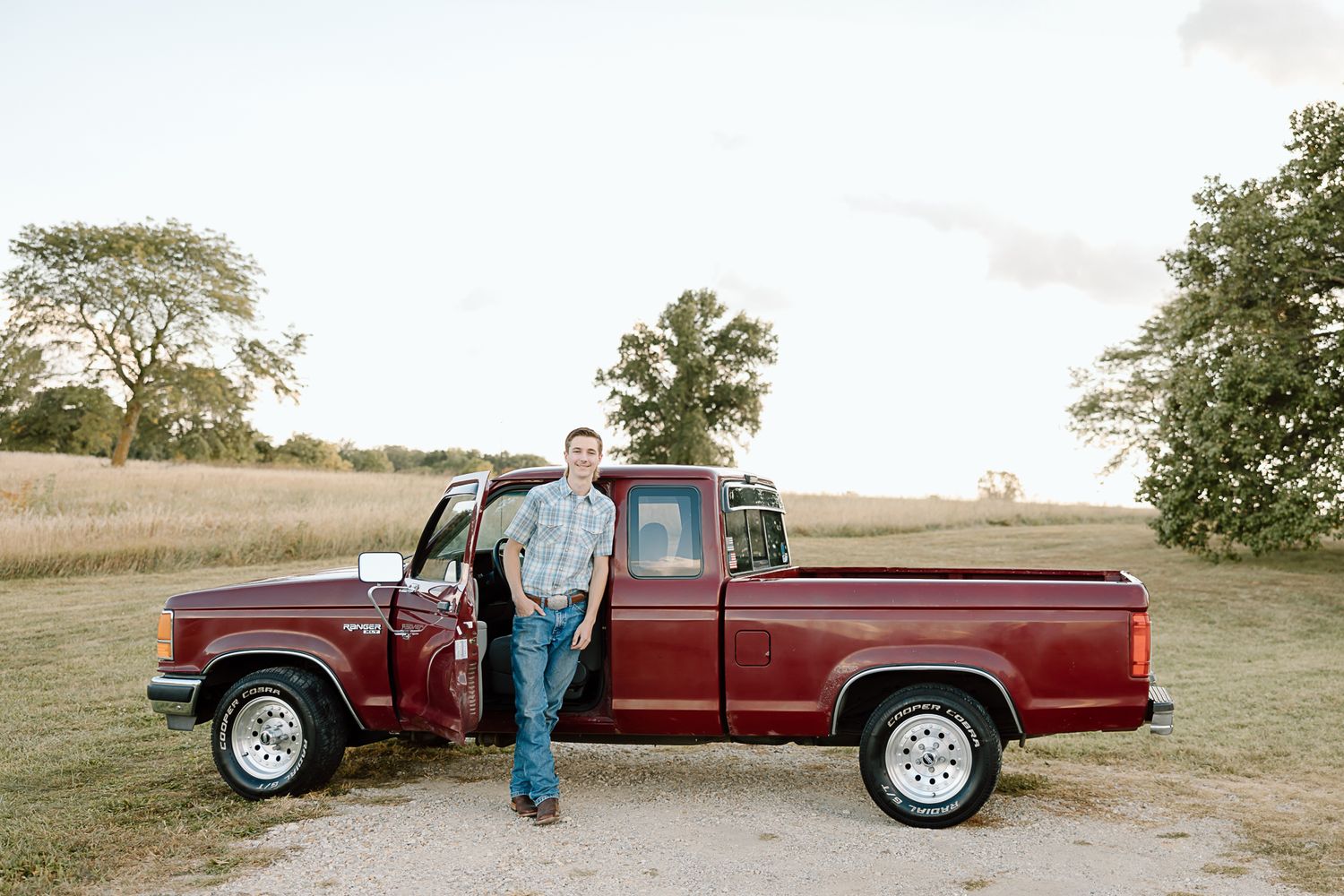 A full-length view of a classic red Ford pickup truck with someone standing beside it in a rural field.