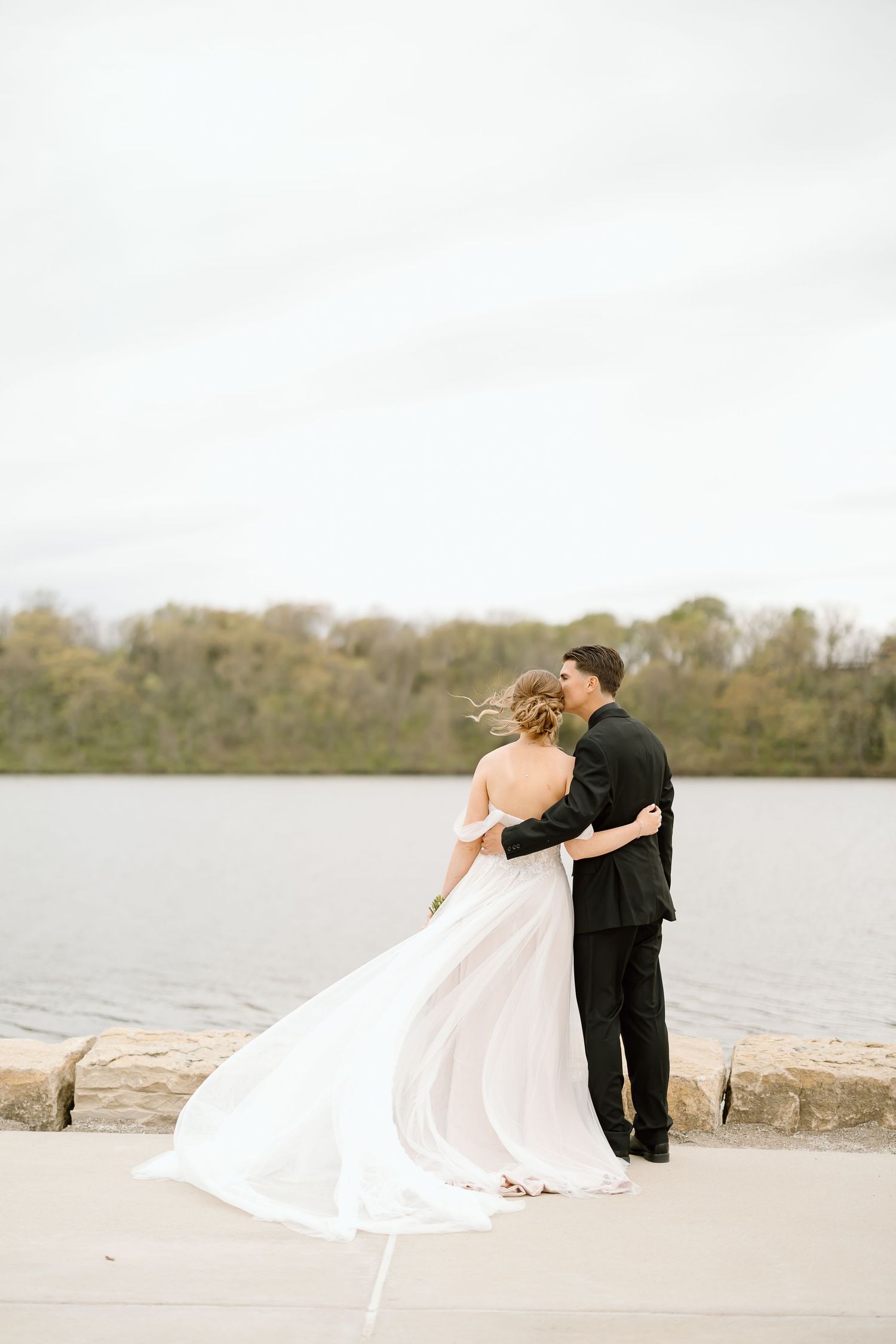 A romantic lakeside moment as newlyweds embrace while standing near stone waterfront.