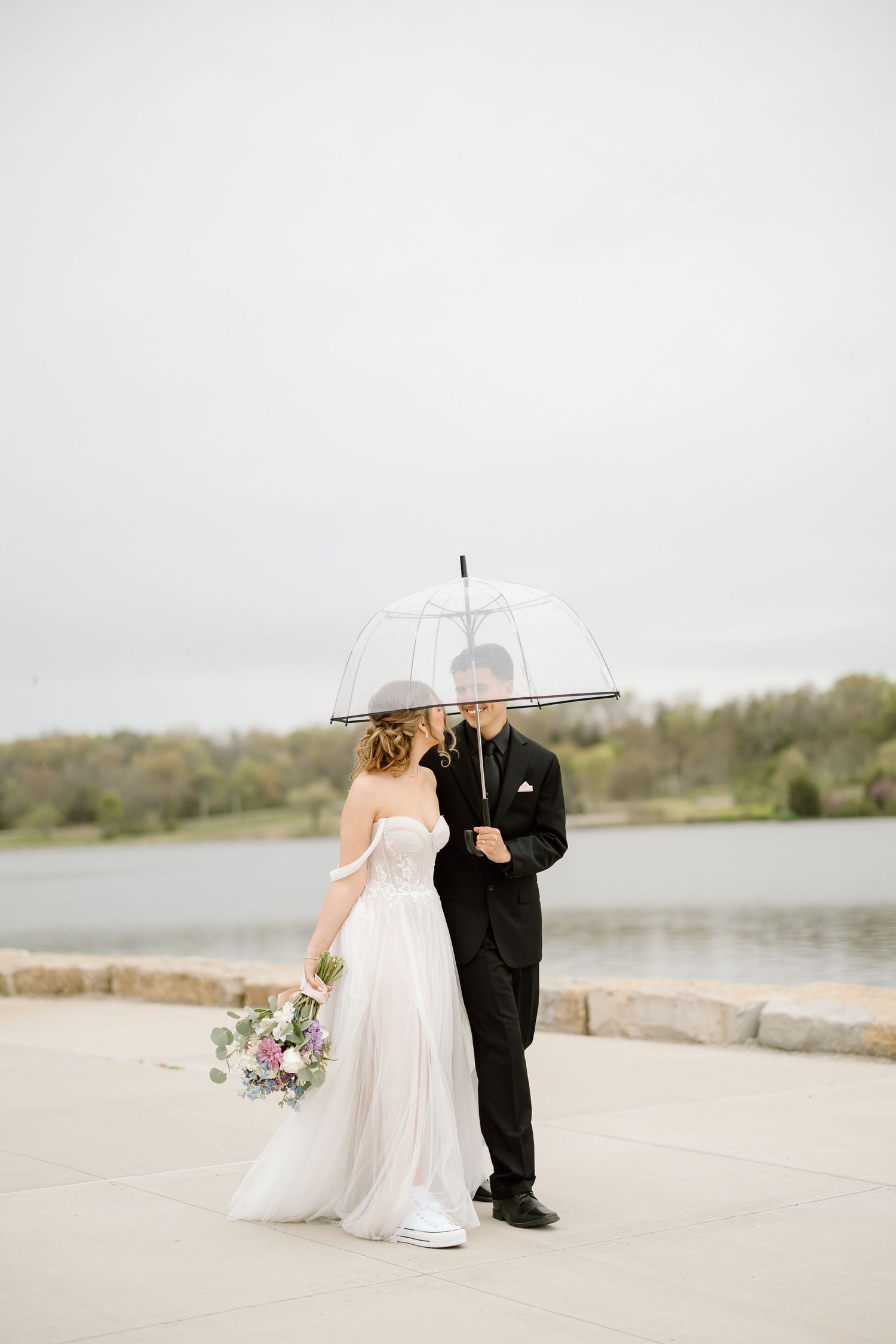 Bride and groom embrace under transparent umbrella beside lake on cloudy day.