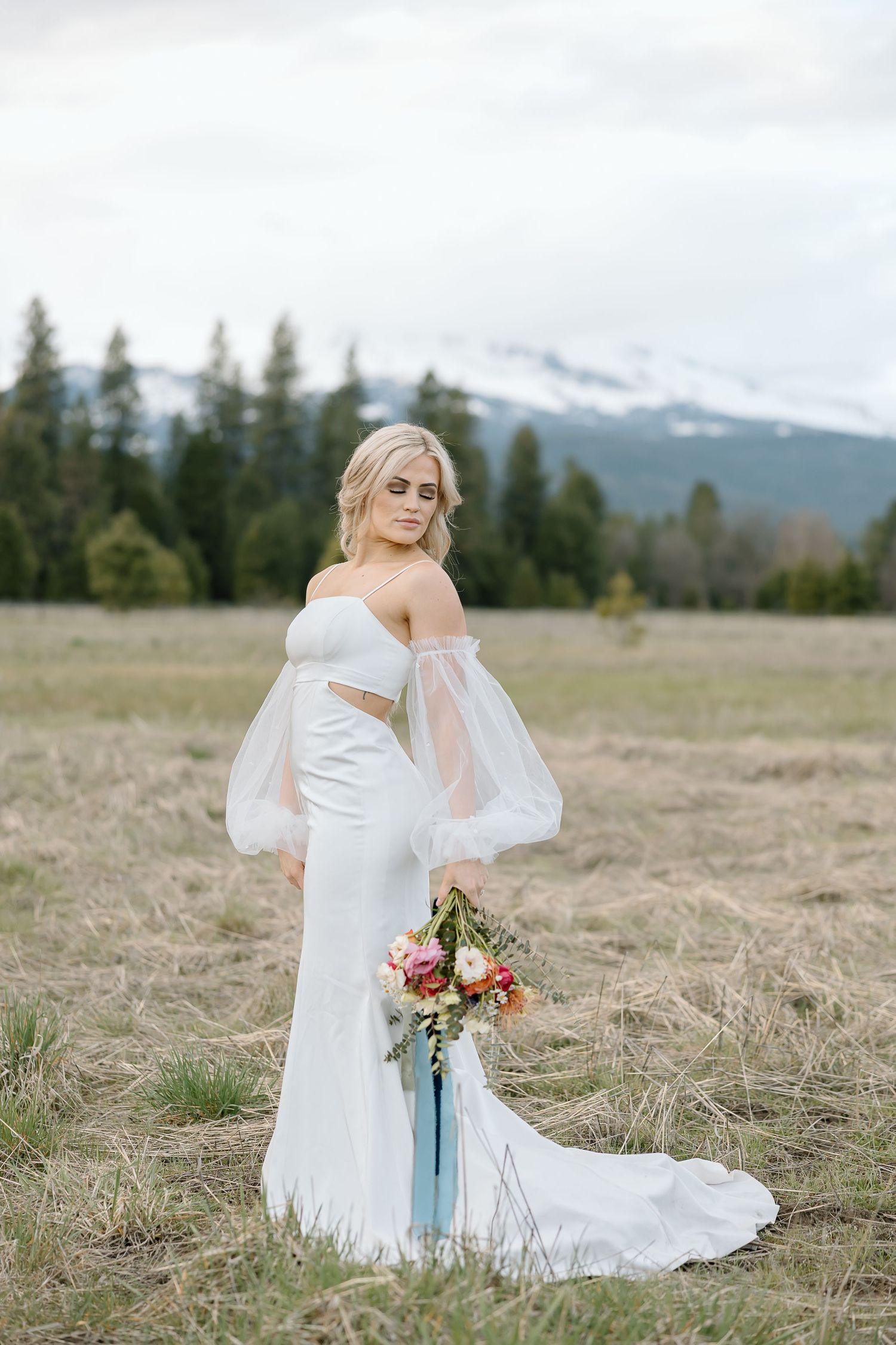 Bride in off-shoulder gown holds colorful bouquet against mountain landscape.