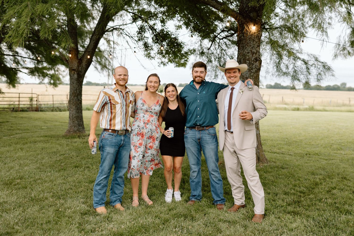 Five friends pose together on a green lawn under trees at an outdoor country gathering on a warm summer evening.