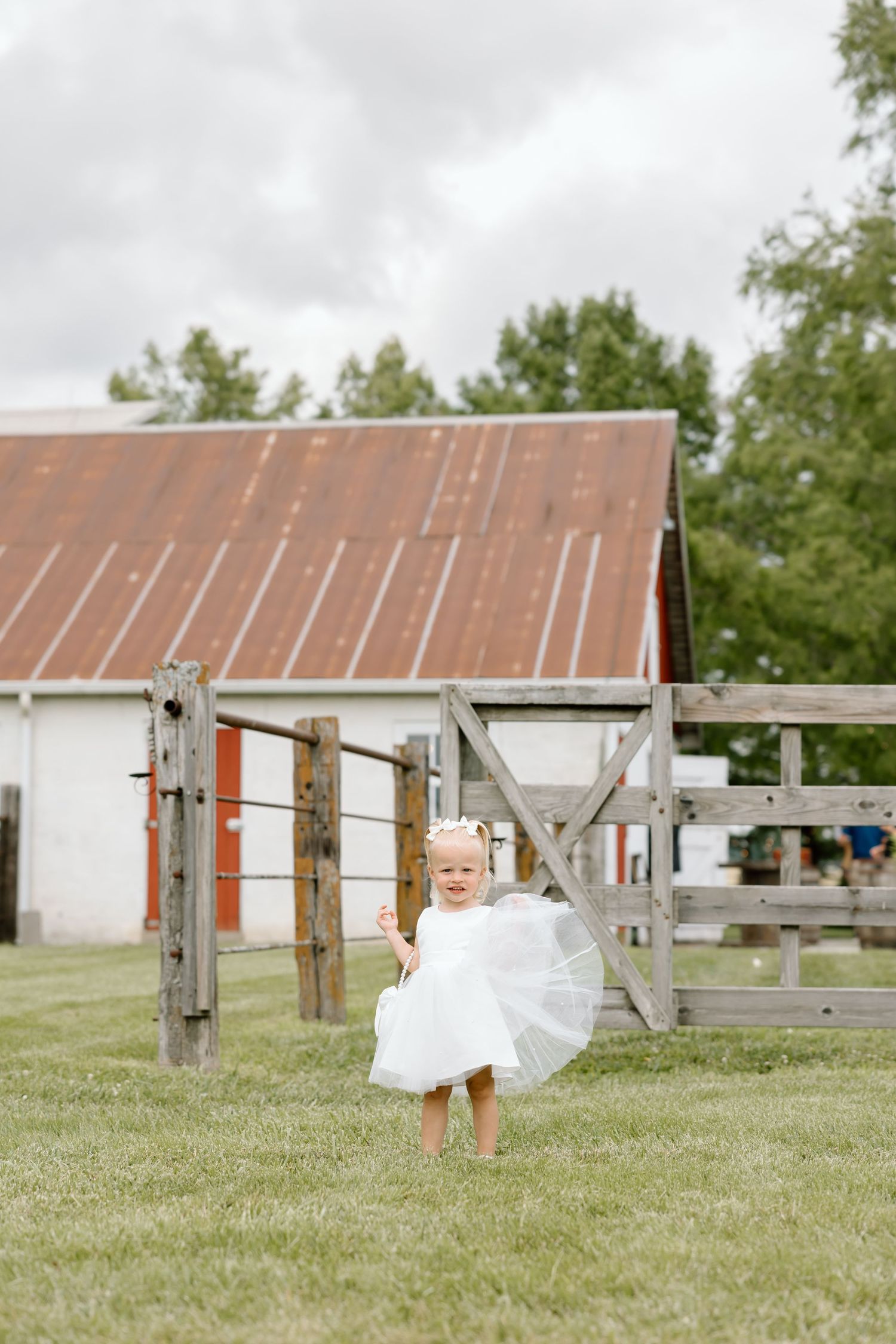 A toddler girl in a white dress holds a small umbrella while standing on grass near a rustic wooden fence and red barn.