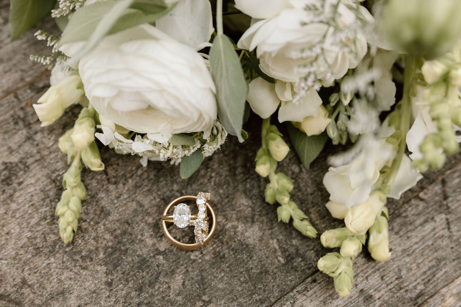 Gold wedding rings resting on rustic wood beside a white floral bouquet with roses and greenery.