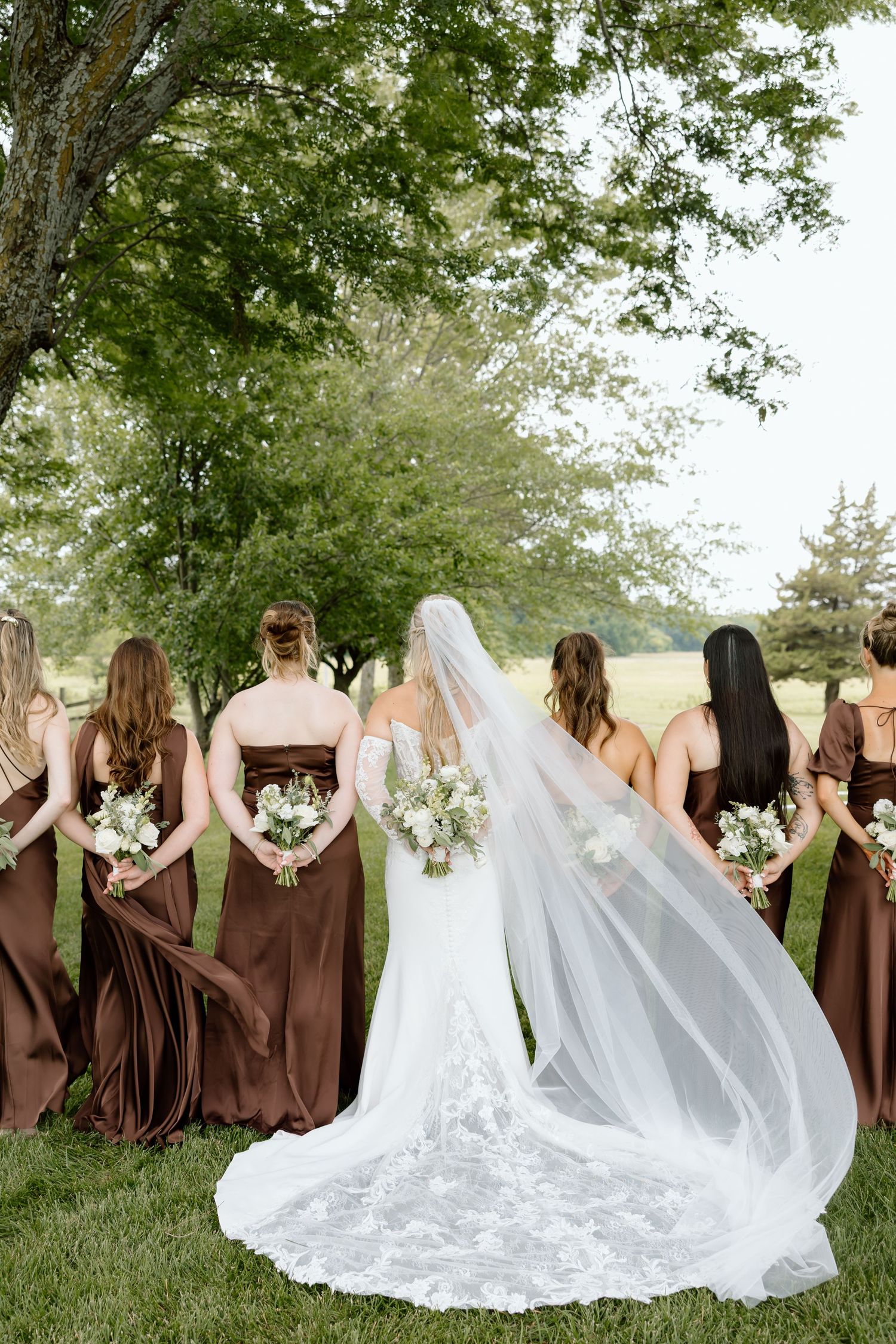 Bride in white lace gown and veil stands with bridesmaids in brown dresses, backs turned, holding bouquets outdoors.